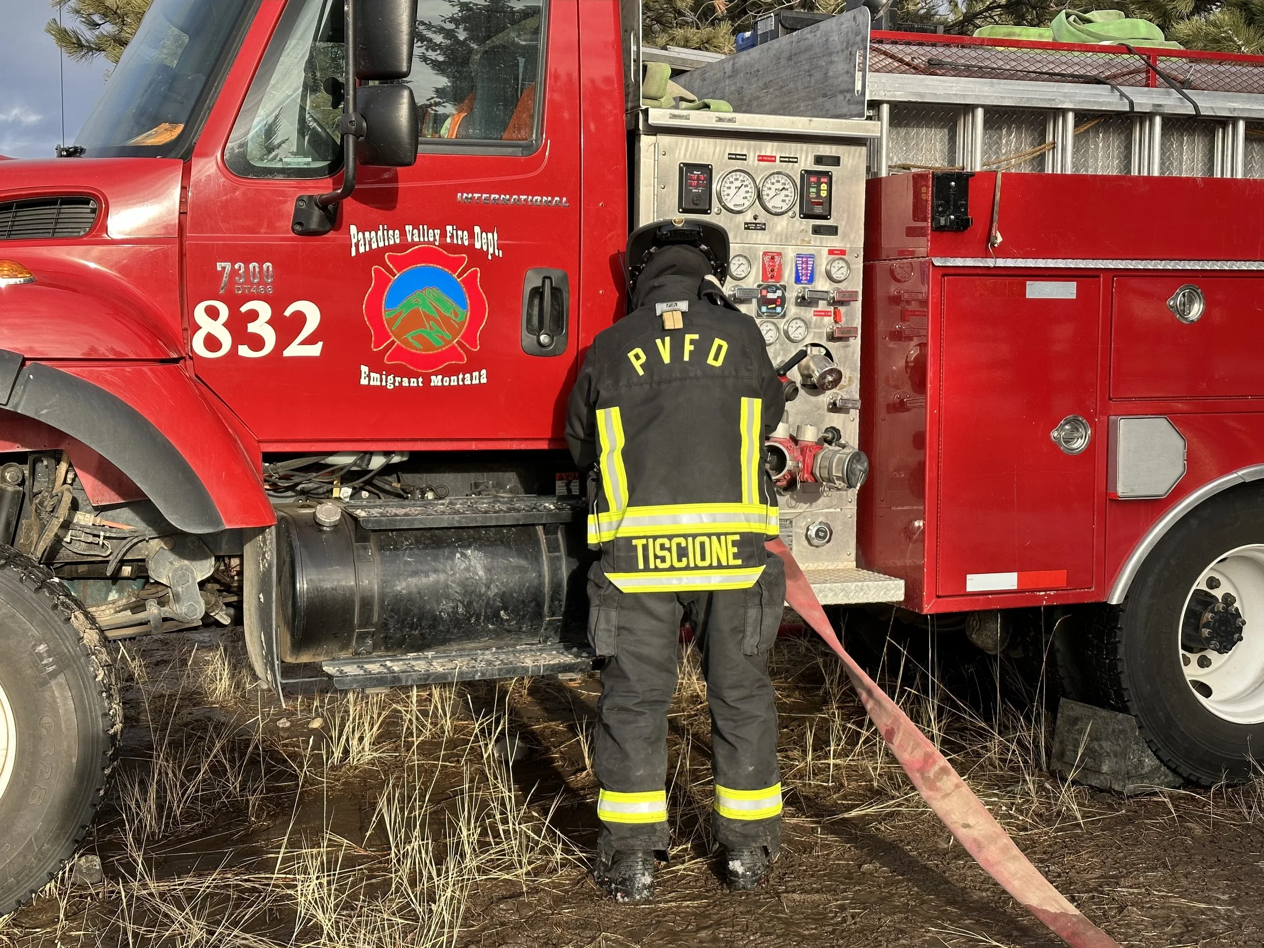 Firefighter operating controls of a red fire truck from Paradise Valley Fire Department, with the firefighter wearing a black uniform with yellow stripes and the text 'PVFD' and 'Tiscione' on the back, in an outdoor setting with grassy ground.