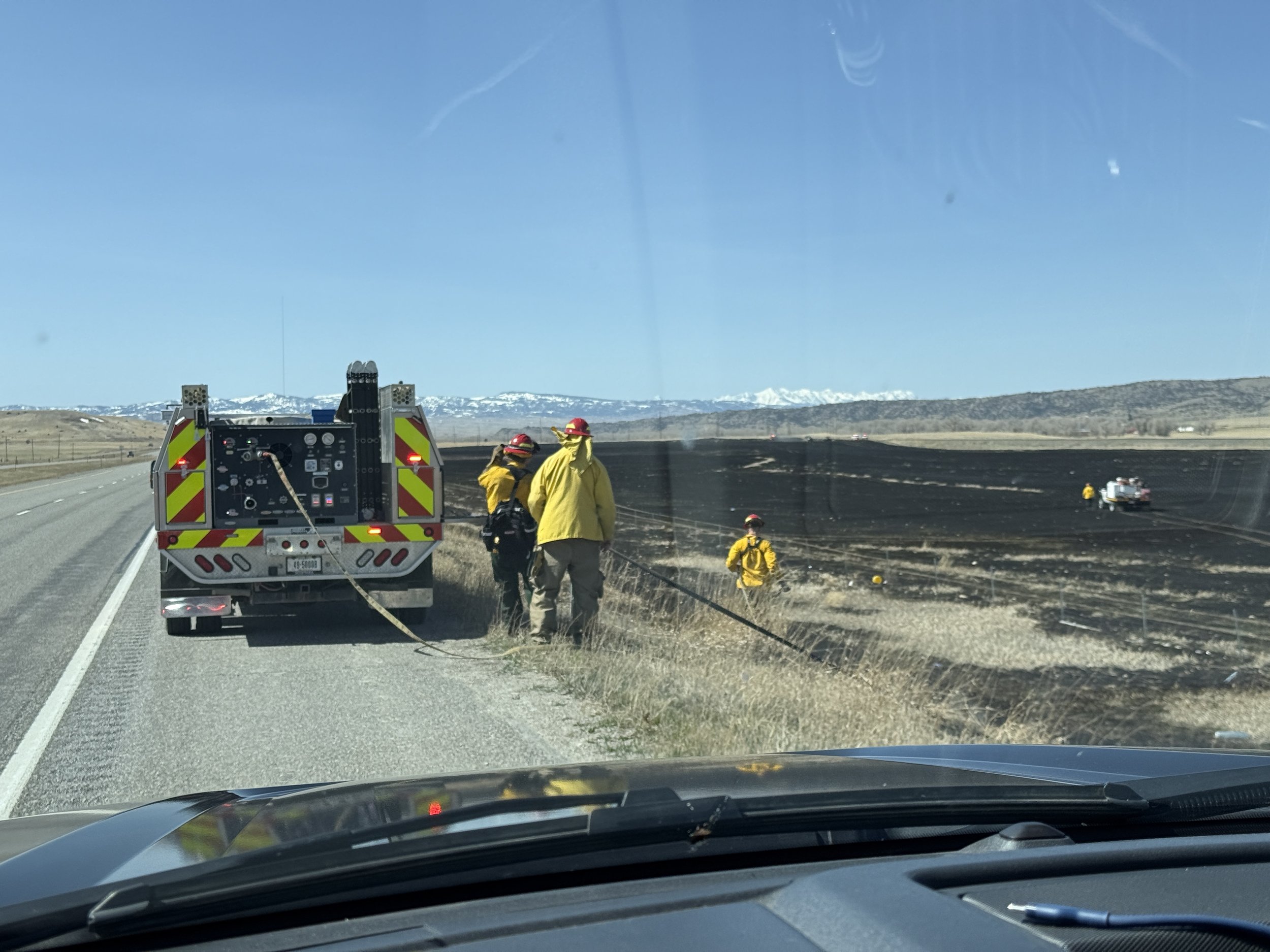 Firefighters and emergency personnel on the side of a highway next to a burn field with blackened soil, working in a rural area with mountains in the distance and a clear blue sky.