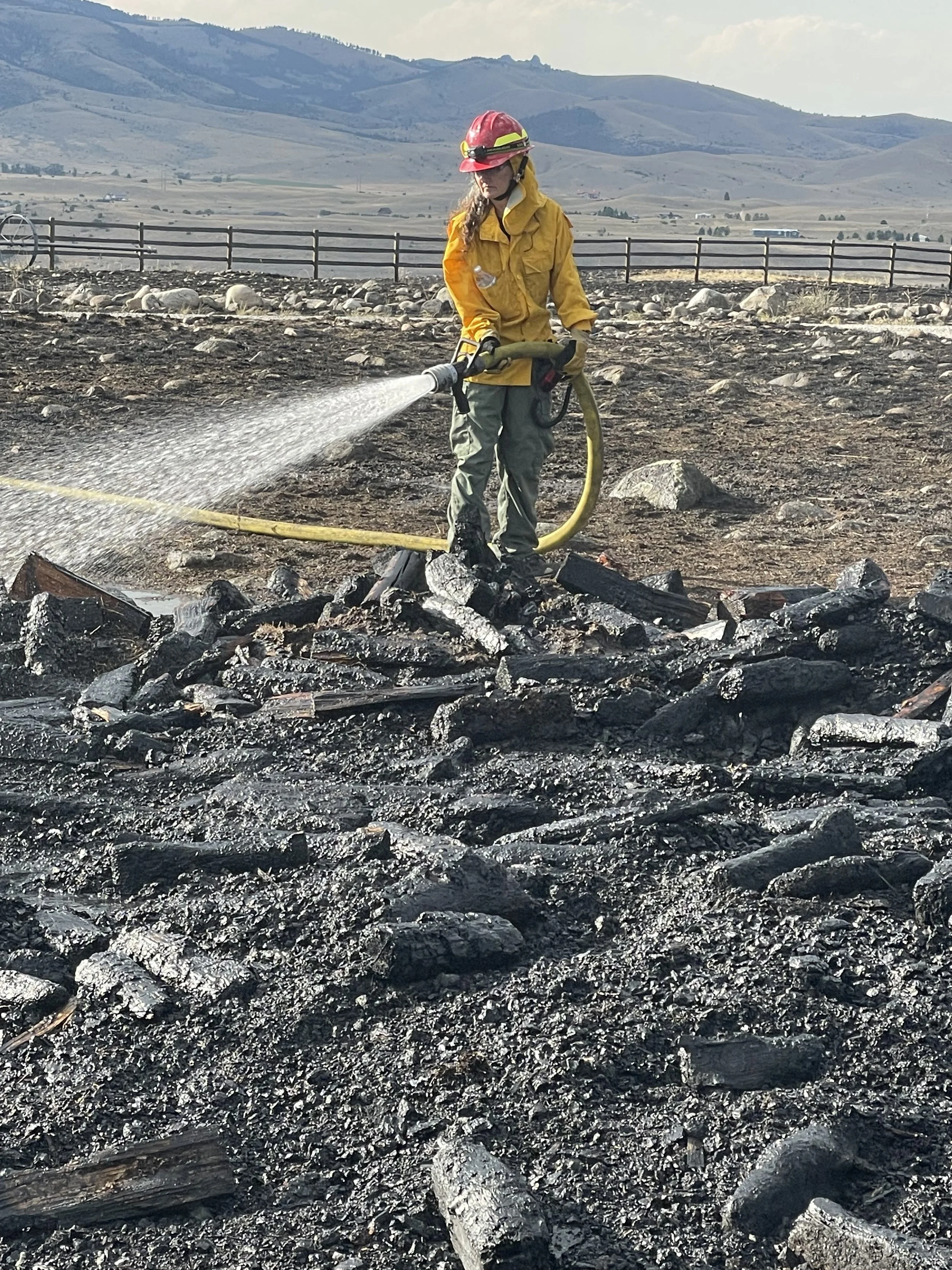 A firefighter spraying water on burnt debris in a wildfire aftermath area near a fence and mountains in the background.