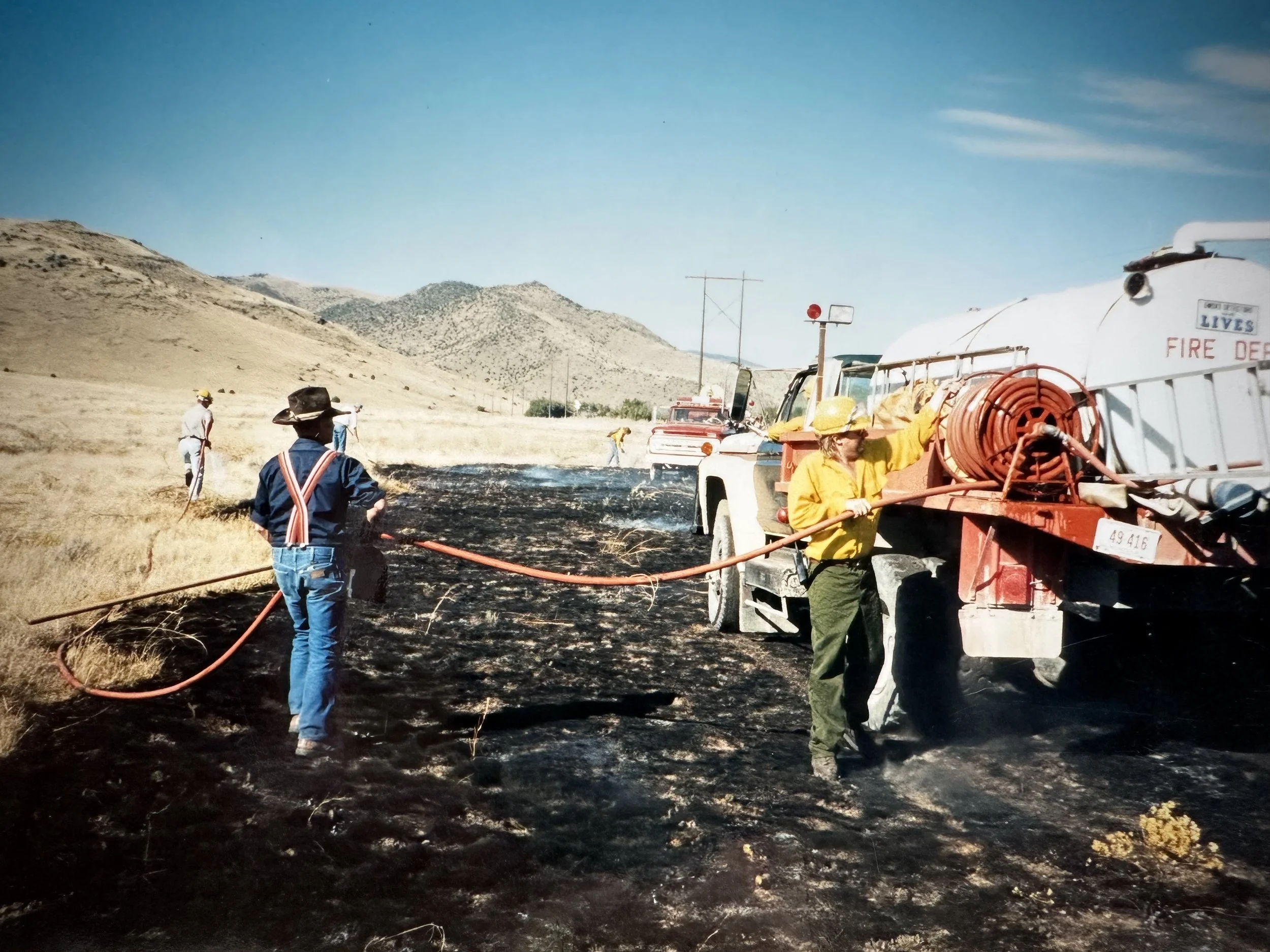 Firefighters and firefighters working with fire hoses near a truck during a wildfire in a dry, hilly landscape under a blue sky.