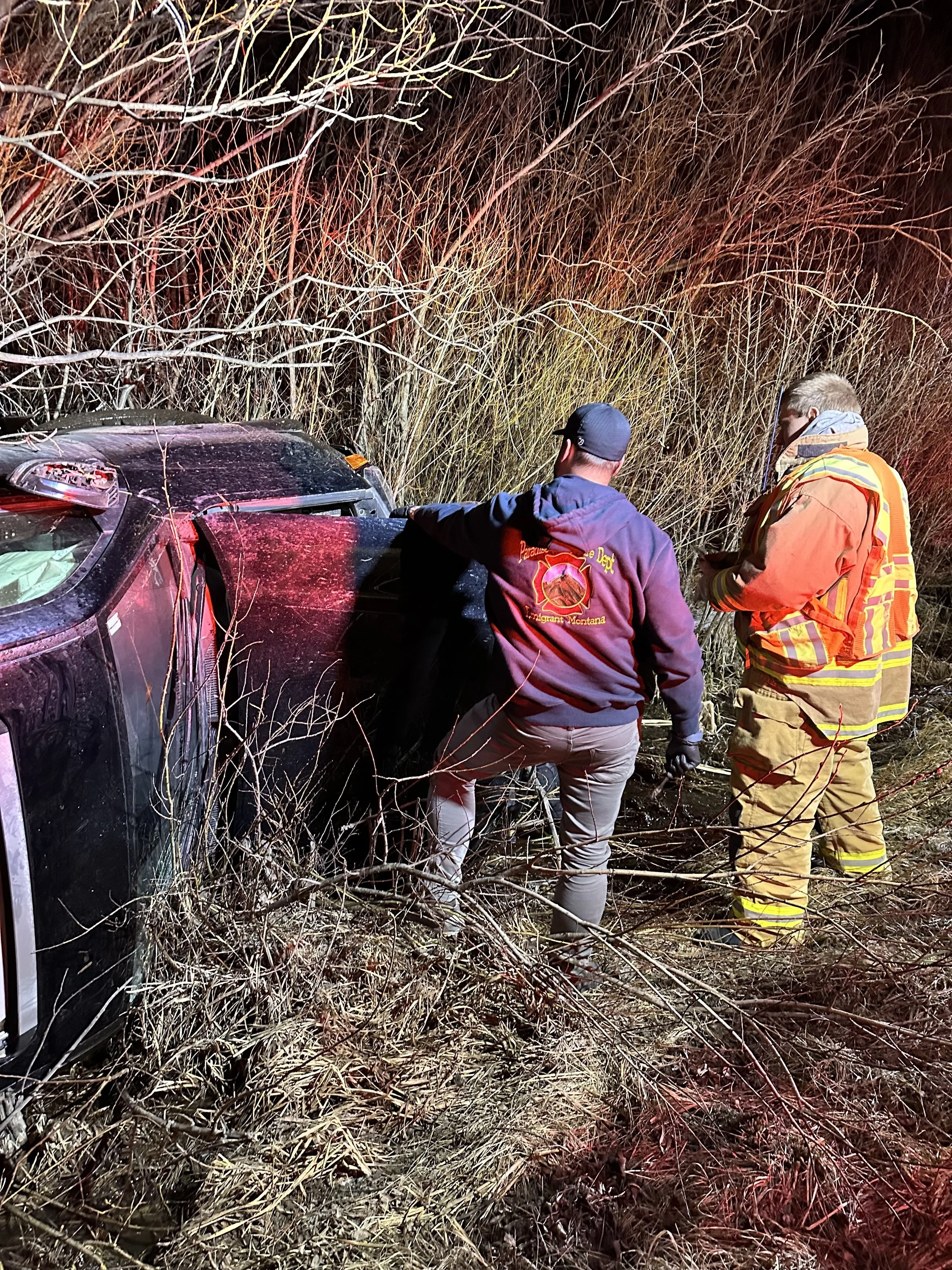 Rescue personnel inspecting a car flipped on its side in a wooded area at night.