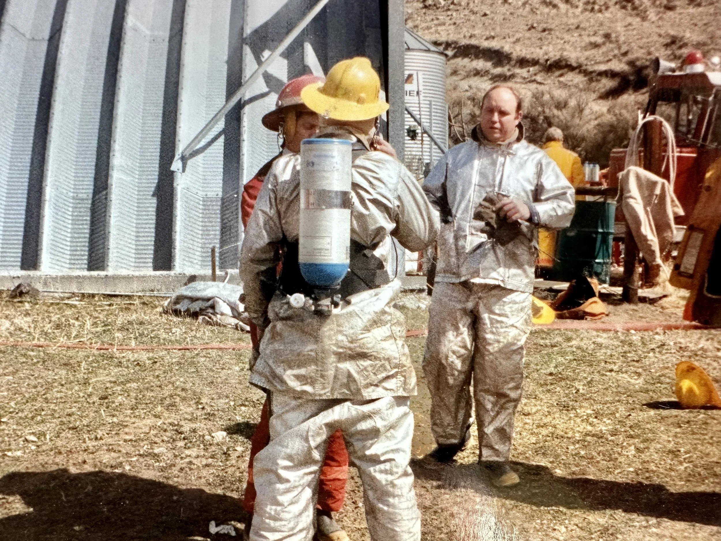 Group of firefighters in silver protective gear and helmets, one with an oxygen tank, standing outdoors near a metal building and machinery, during a fire drill or emergency.