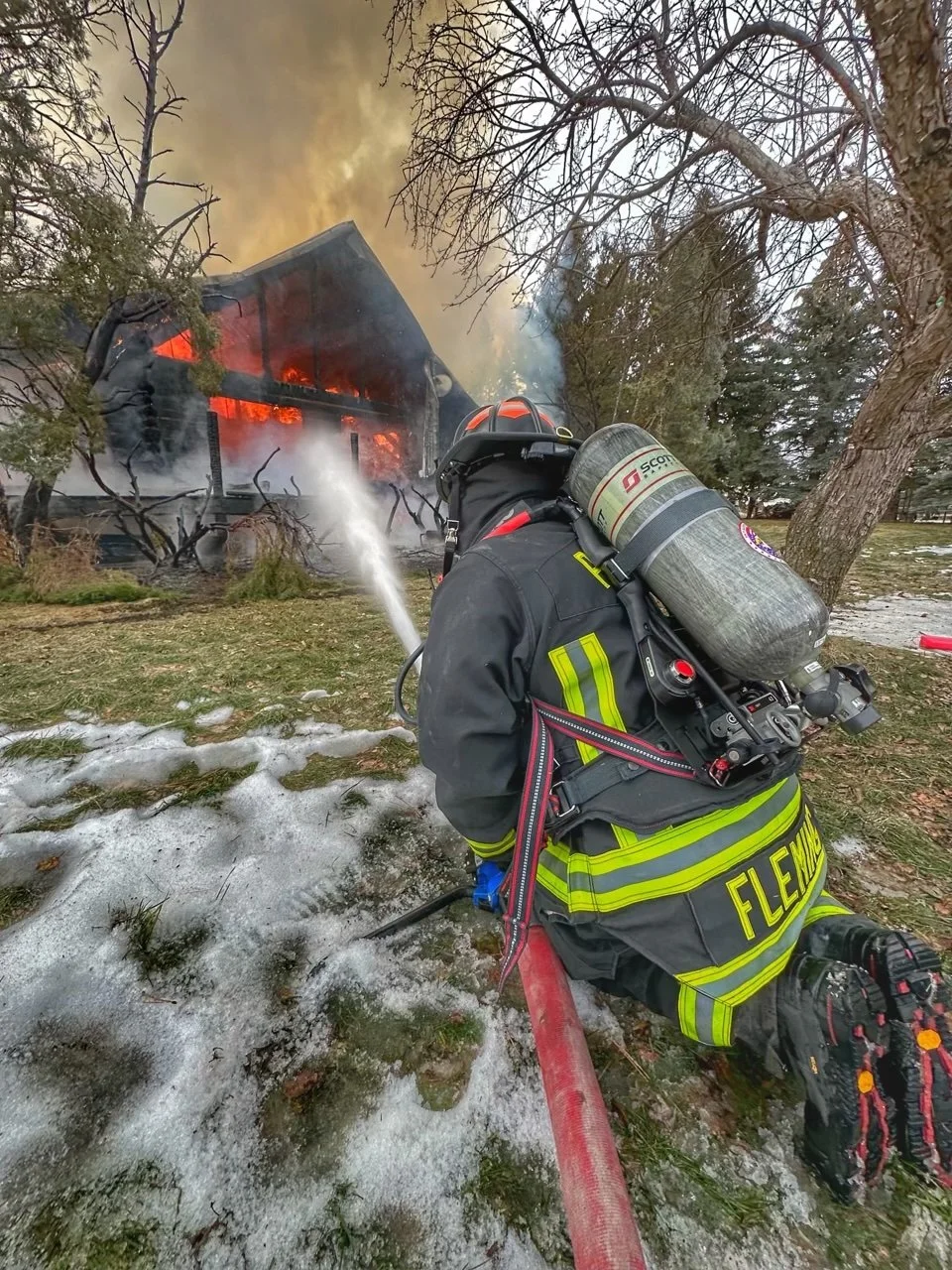 A firefighter in full gear, including a breathing tank, is kneeling on the ground and spraying water from a hose at a house that is on fire with flames and smoke coming out of the windows and roof.
