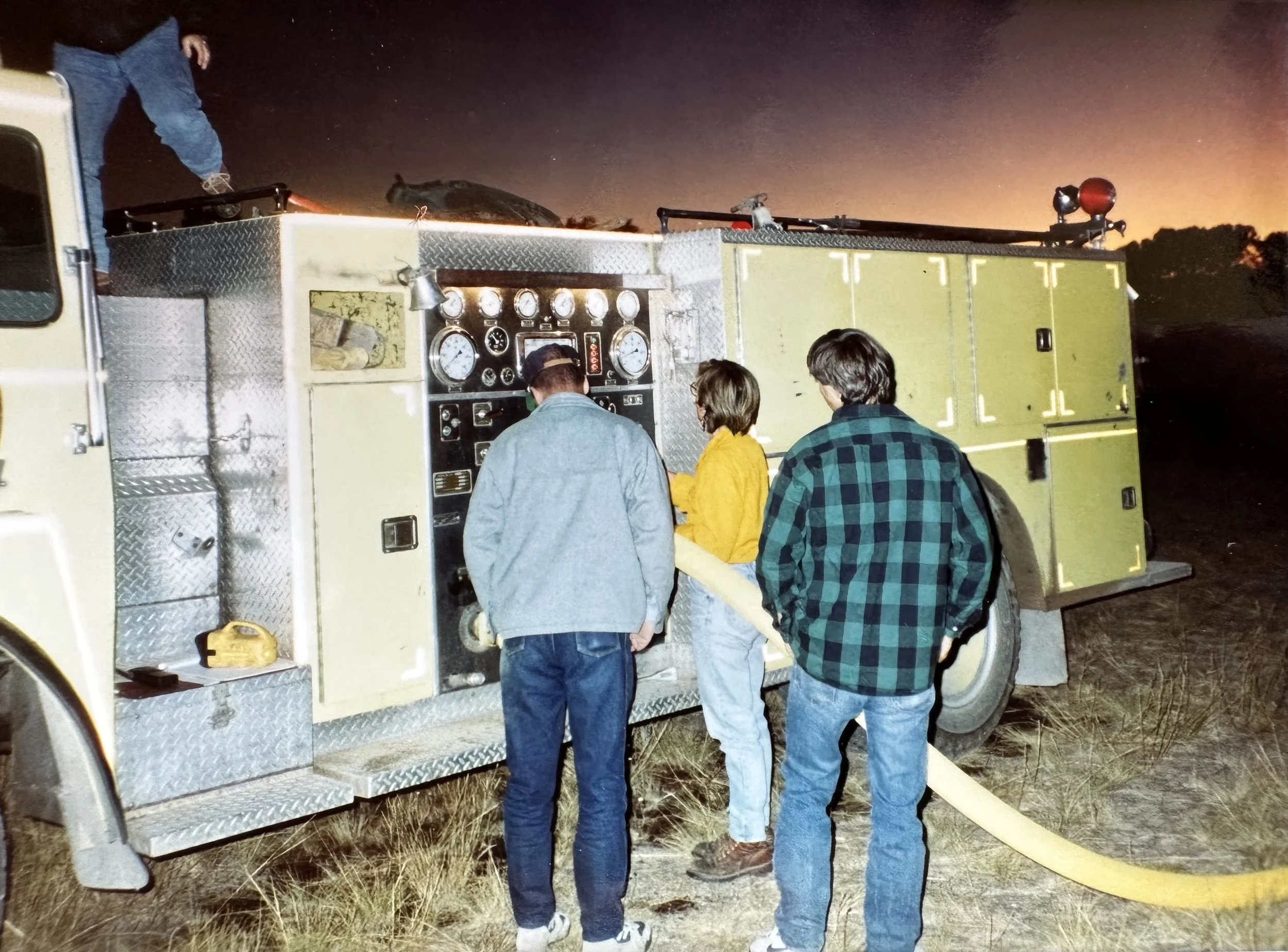 Three people, two men and one woman, are gathered around the control panel of a fire truck, which is parked outdoors at dusk. The woman is wearing a yellow jacket, and the men are wearing plaid and denim jackets. The scene has a rural or open field b