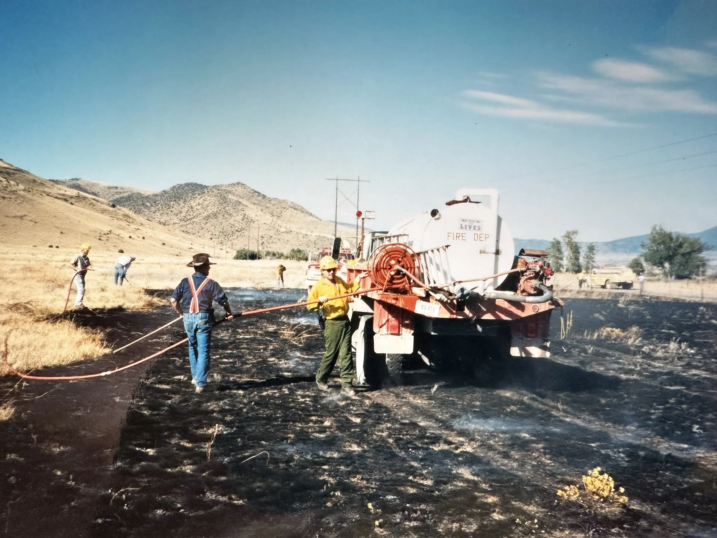 Firefighters and workers extinguishing a grass fire in a rural area with mountains in the background. Fire truck labeled 'Living Lives Fire Dept' is spraying water on the burned grass.