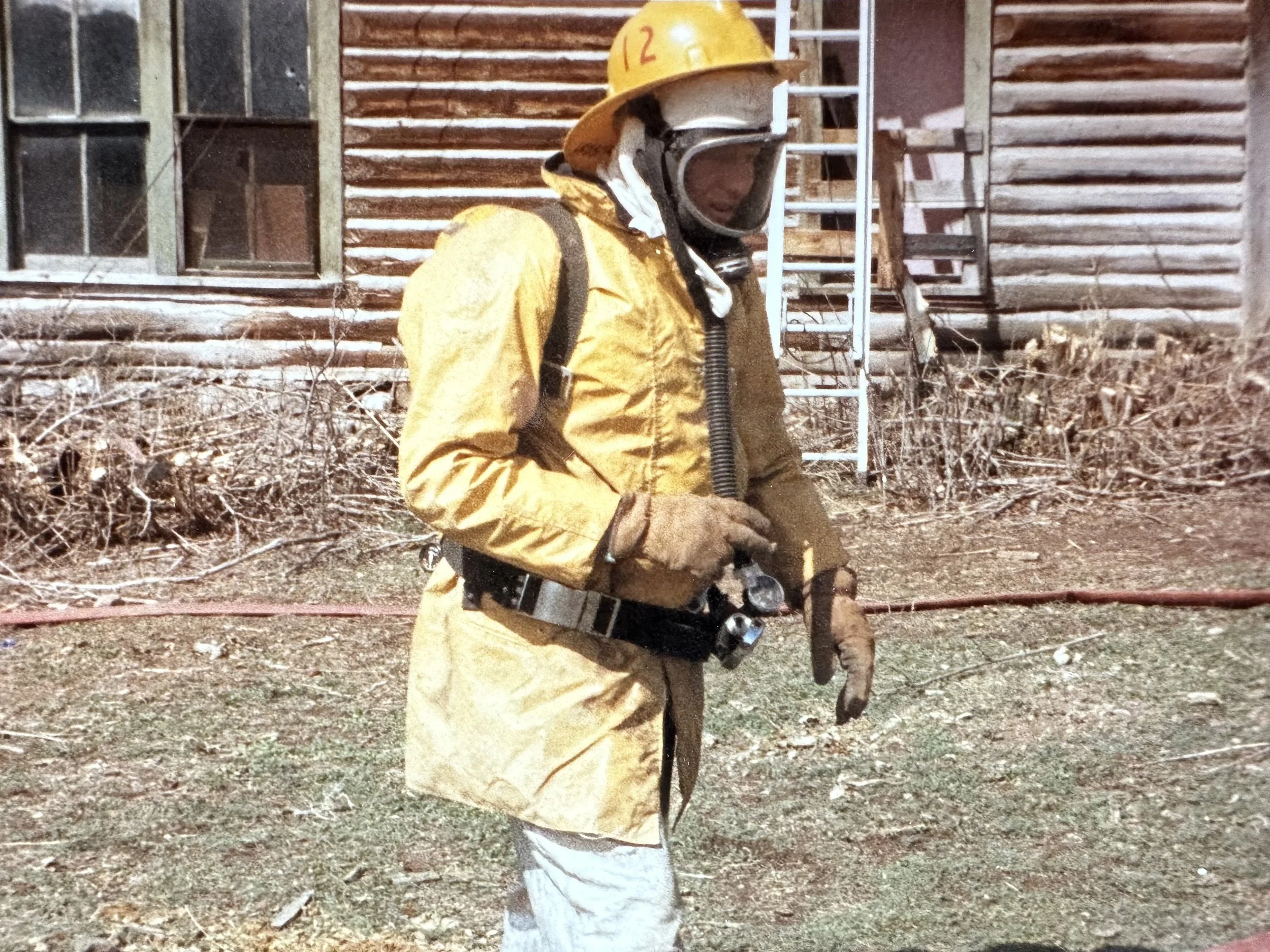 Person dressed in yellow firefighting suit with helmet, gloves, and breathing mask, standing outdoors near a wooden house with ladder and brush in the background.