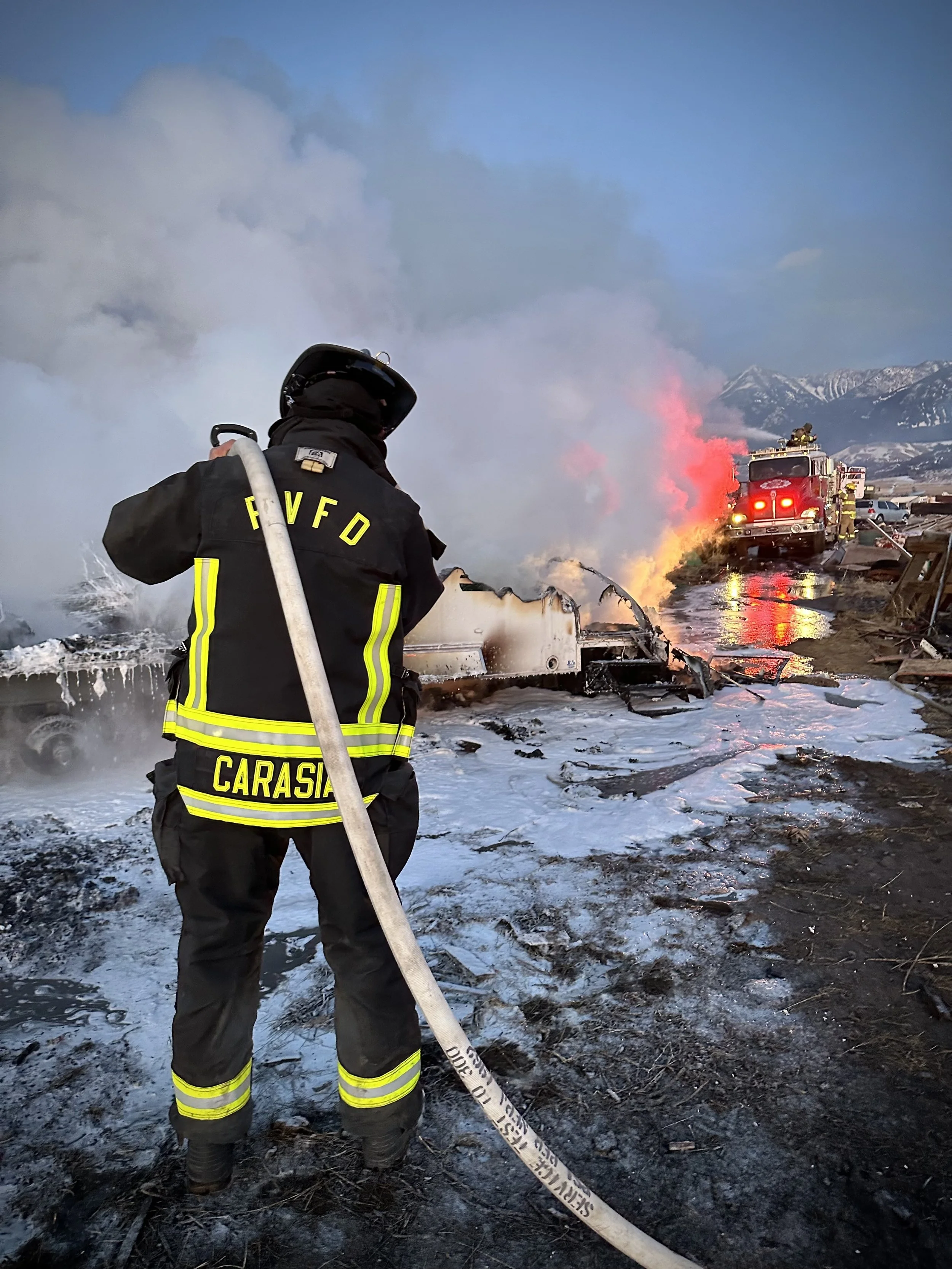 Firefighter in firefighting gear is spraying water on a burned vehicle at a fire scene with smoke and flames, alongside other fire trucks, mountains in the background.