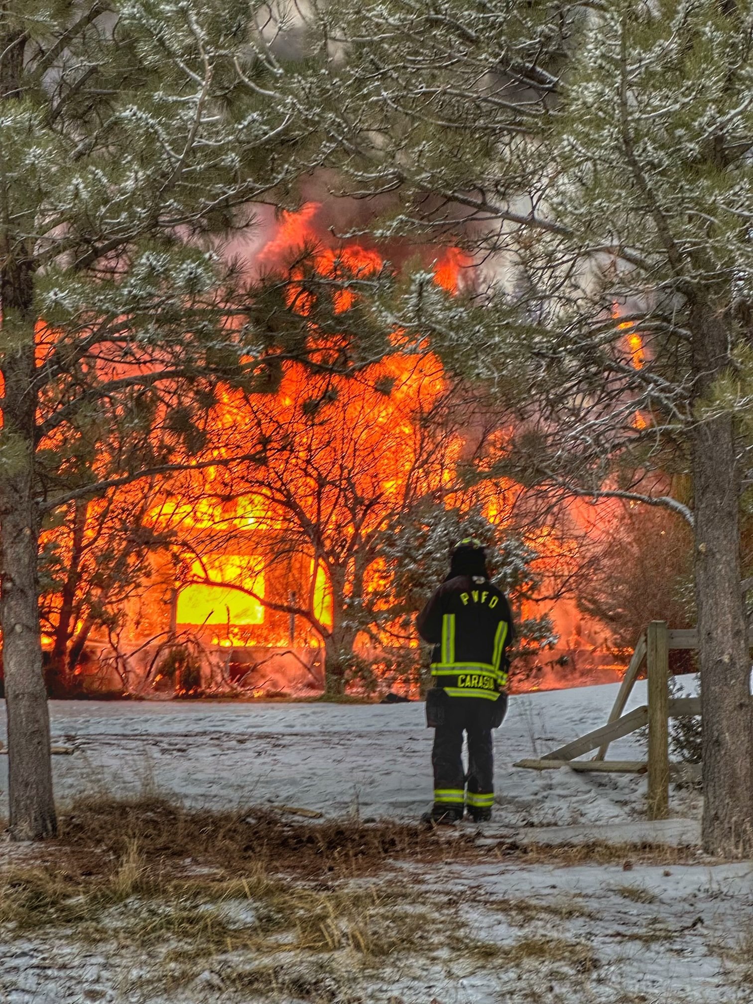 Firefighter standing on snow-covered ground watching a large house fire with flames and smoke visible through trees.