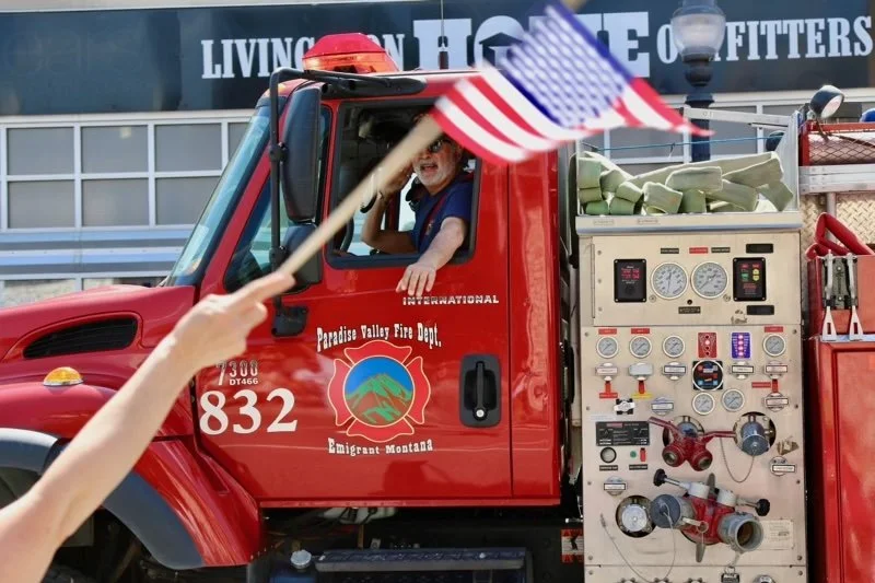 A person waving an American flag in front of a fire truck from Paradise Valley Fire Department, with a firefighter's station in the background.