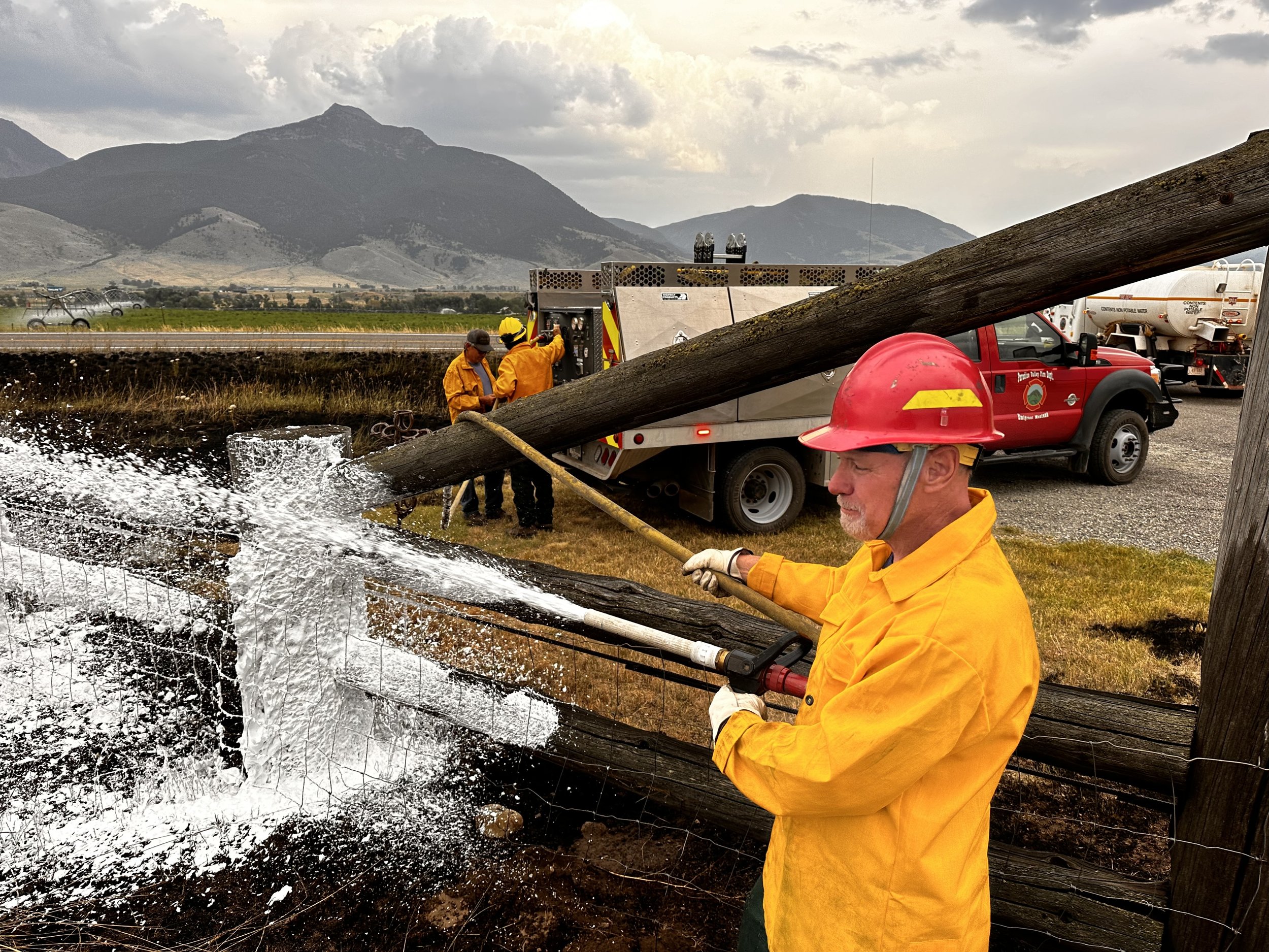Firefighters working on a toppled utility pole, spraying water to stabilize or remove it, with mountains and cloudy sky in the background.