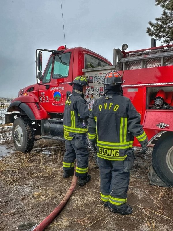 Two firefighters in black gear with reflective yellow stripes and helmets stand next to a red fire truck on a muddy field with sparse grass, operating controls on the side of the fire truck.