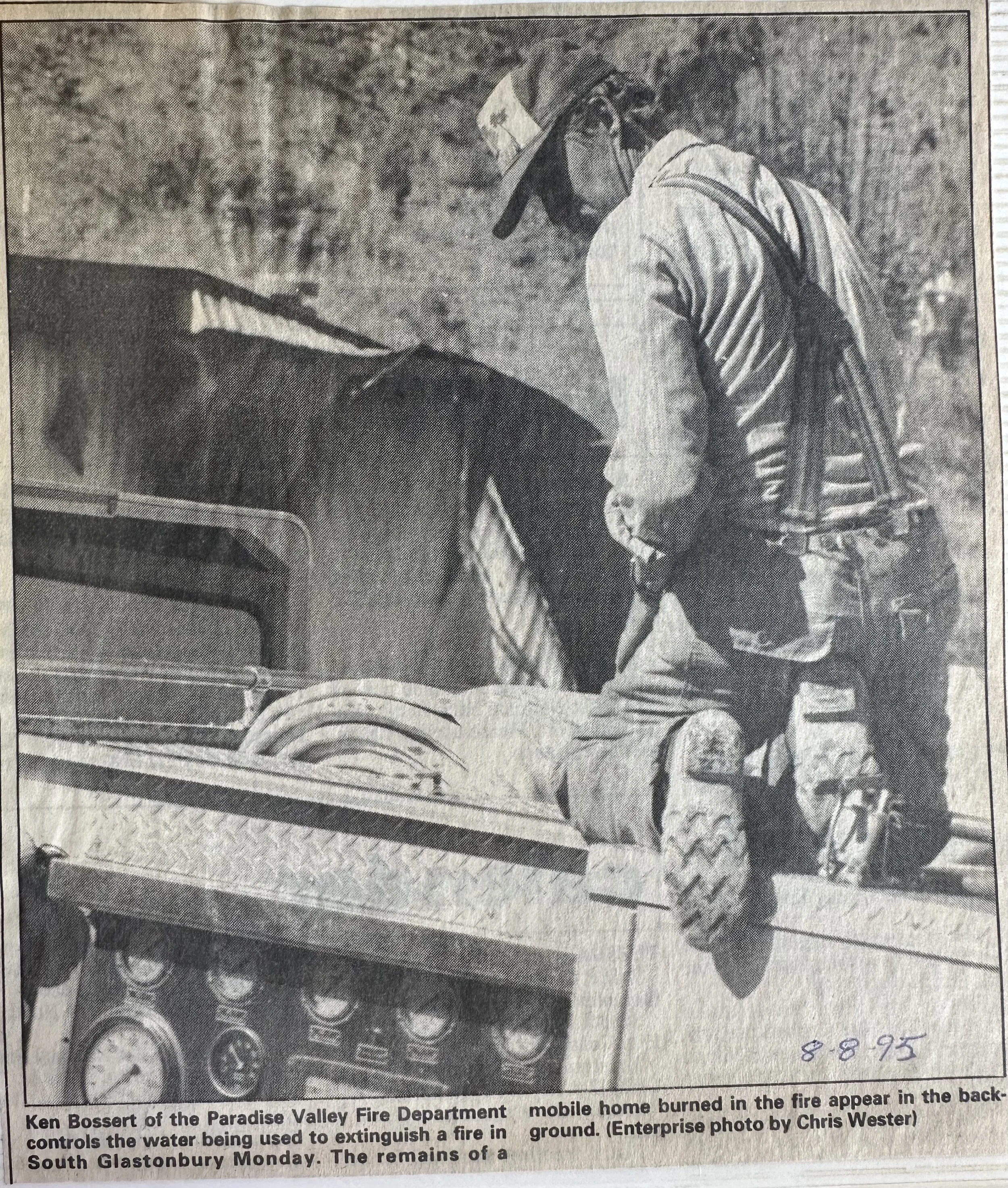 A firefighter in uniform and helmet operating a fire truck's control panel, with a fire burning in the background.