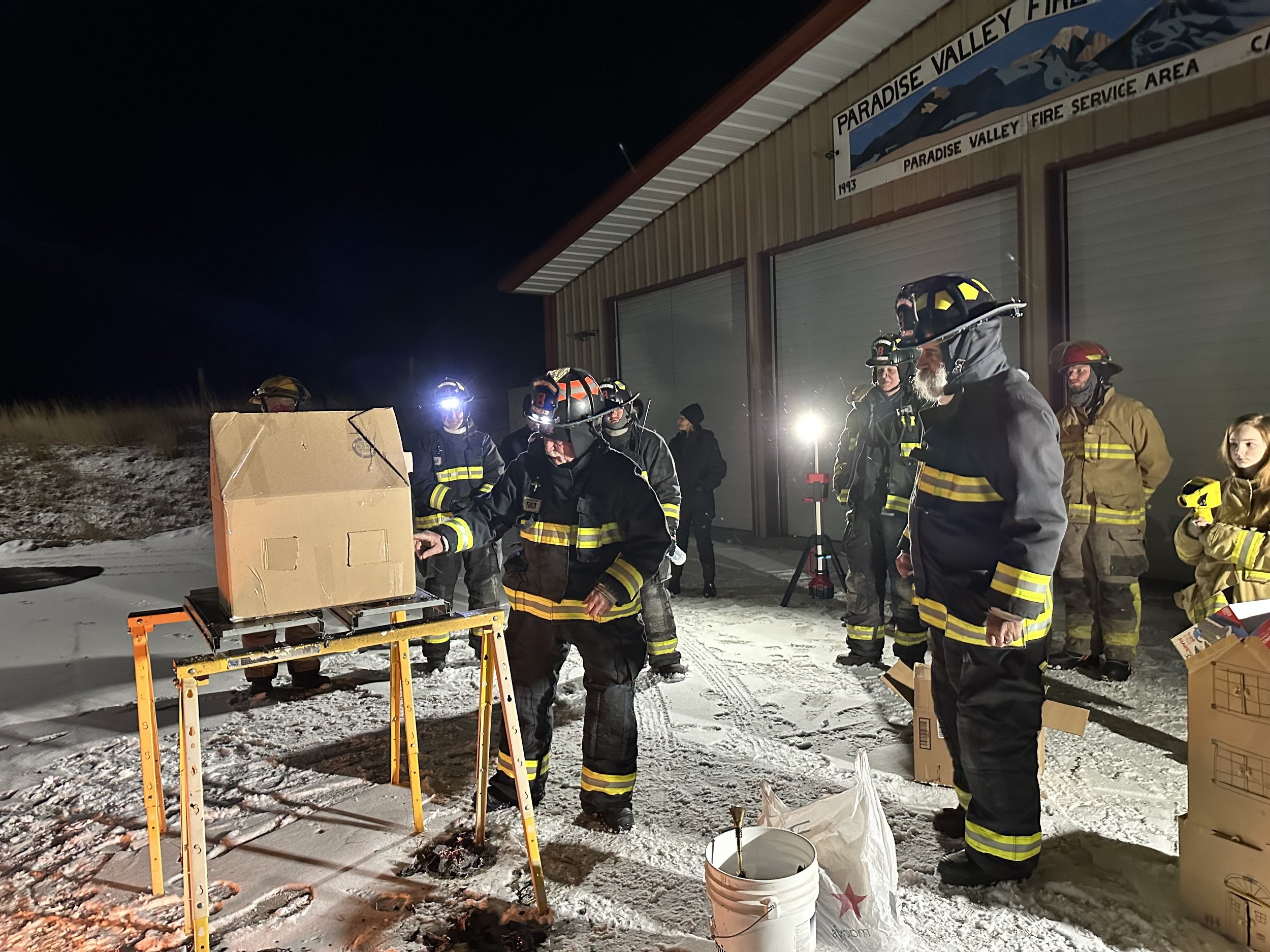 Firefighters conducting a night training exercise with a cardboard box on a table outside a fire station.