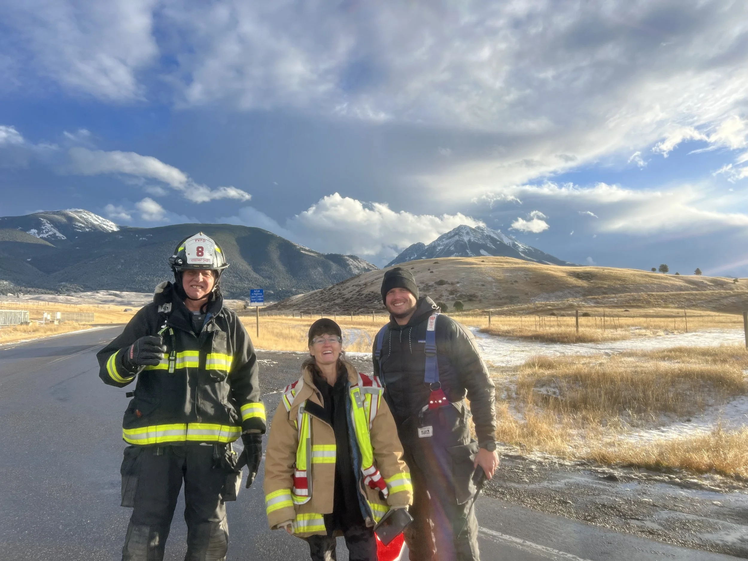 Three emergency responders standing on a road with mountains in the background. The responders are wearing jackets and helmets, smiling, with a partly cloudy sky overhead.