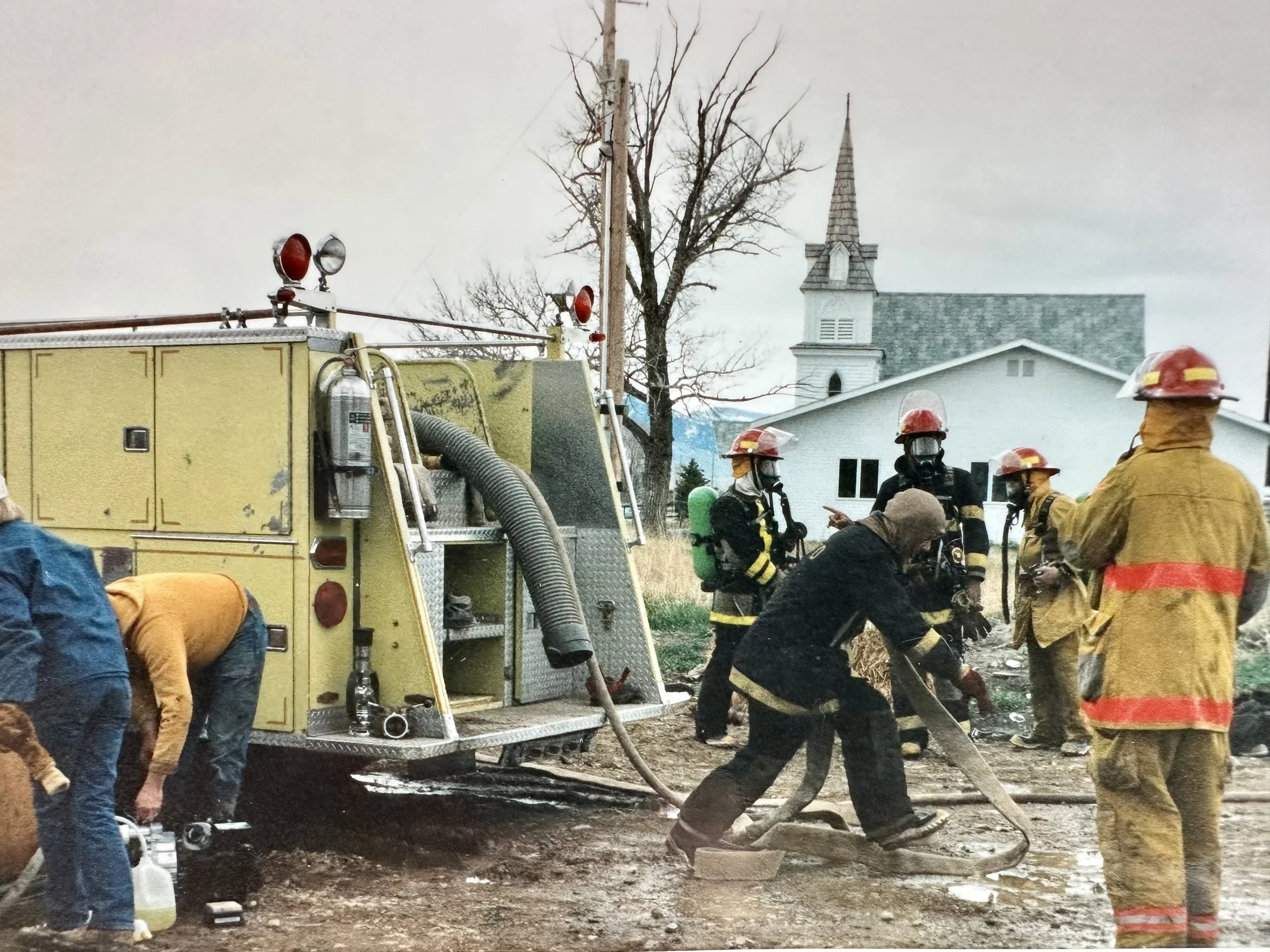 Firefighters and rescue personnel working near a yellow fire truck with hoses, on a muddy street in front of a white church building with a steeple.