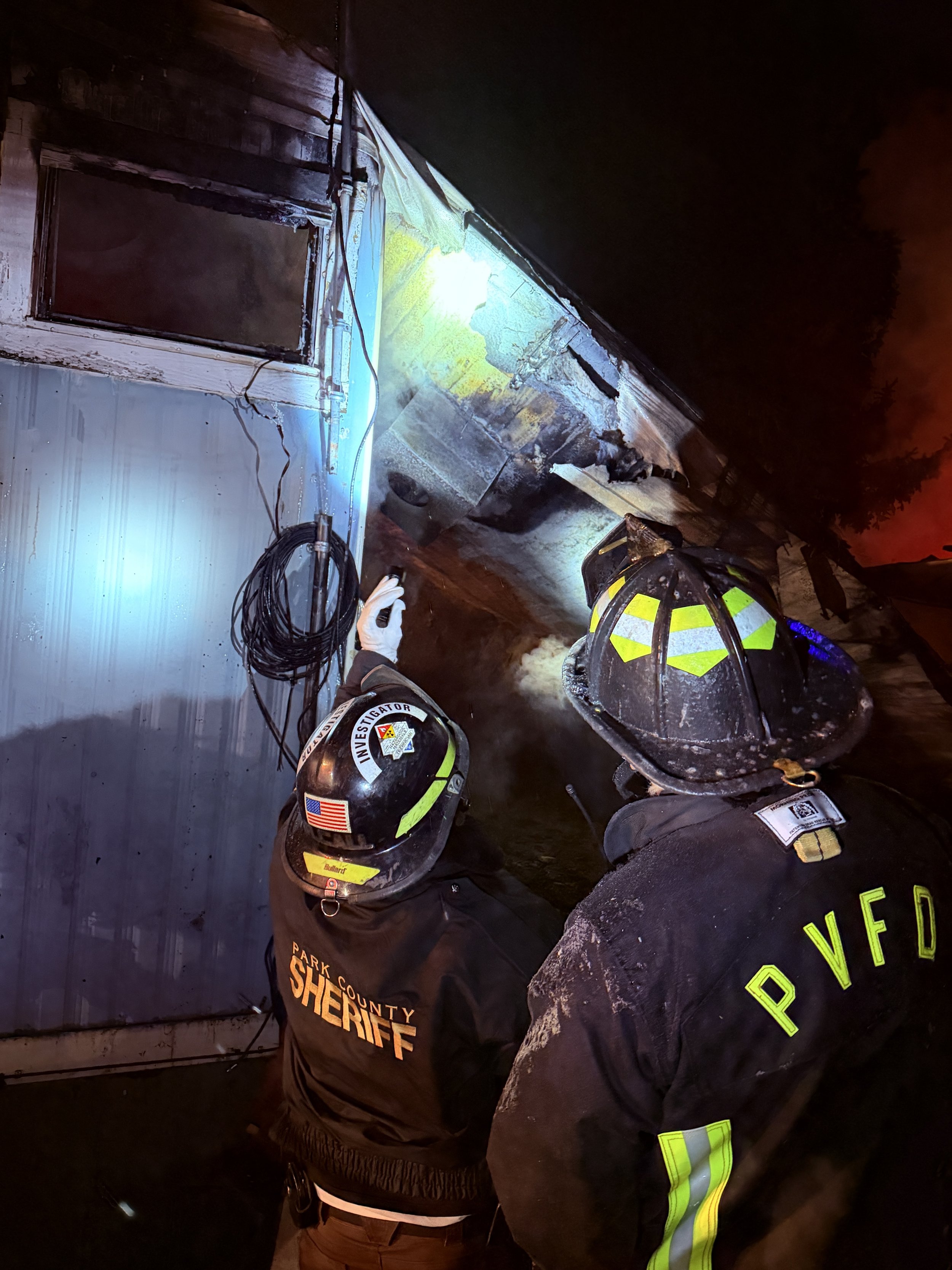 Two firefighters investigating a night-time house fire with smoke and damage visible on the house's exterior wall.