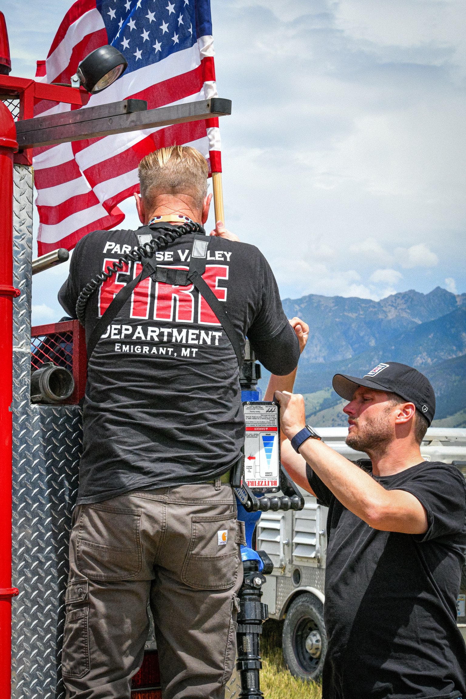 Two firefighters are working on a fire truck in a rural area with mountains in the background. One firefighter, wearing a black shirt with 'Parrosee Valley Fire Department Emigrant, MT' on the back, is standing on the truck, while the other firefight