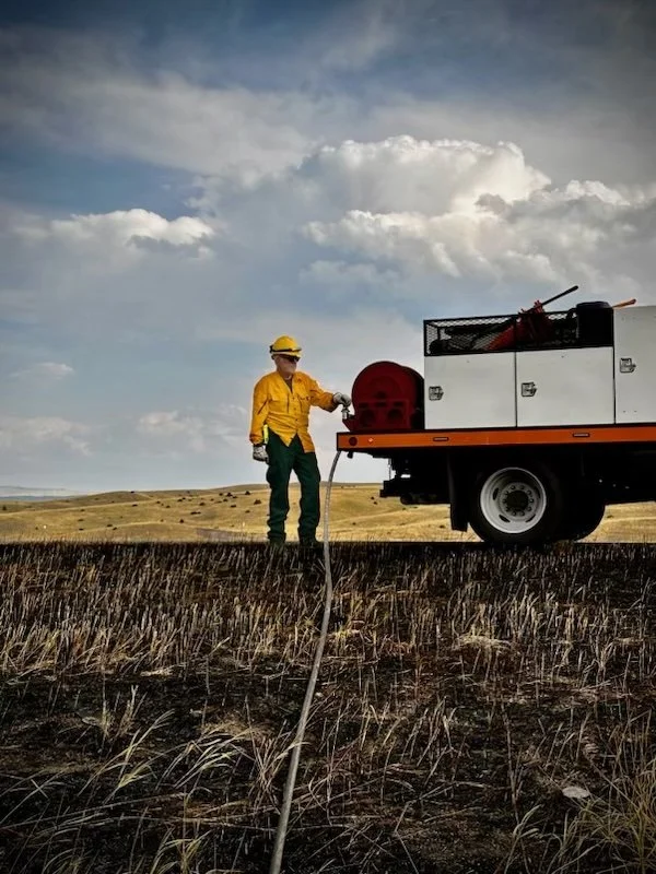 A firefighter in yellow and green gear holding a hose connects to a fire truck in an open field with a cloudy sky above.