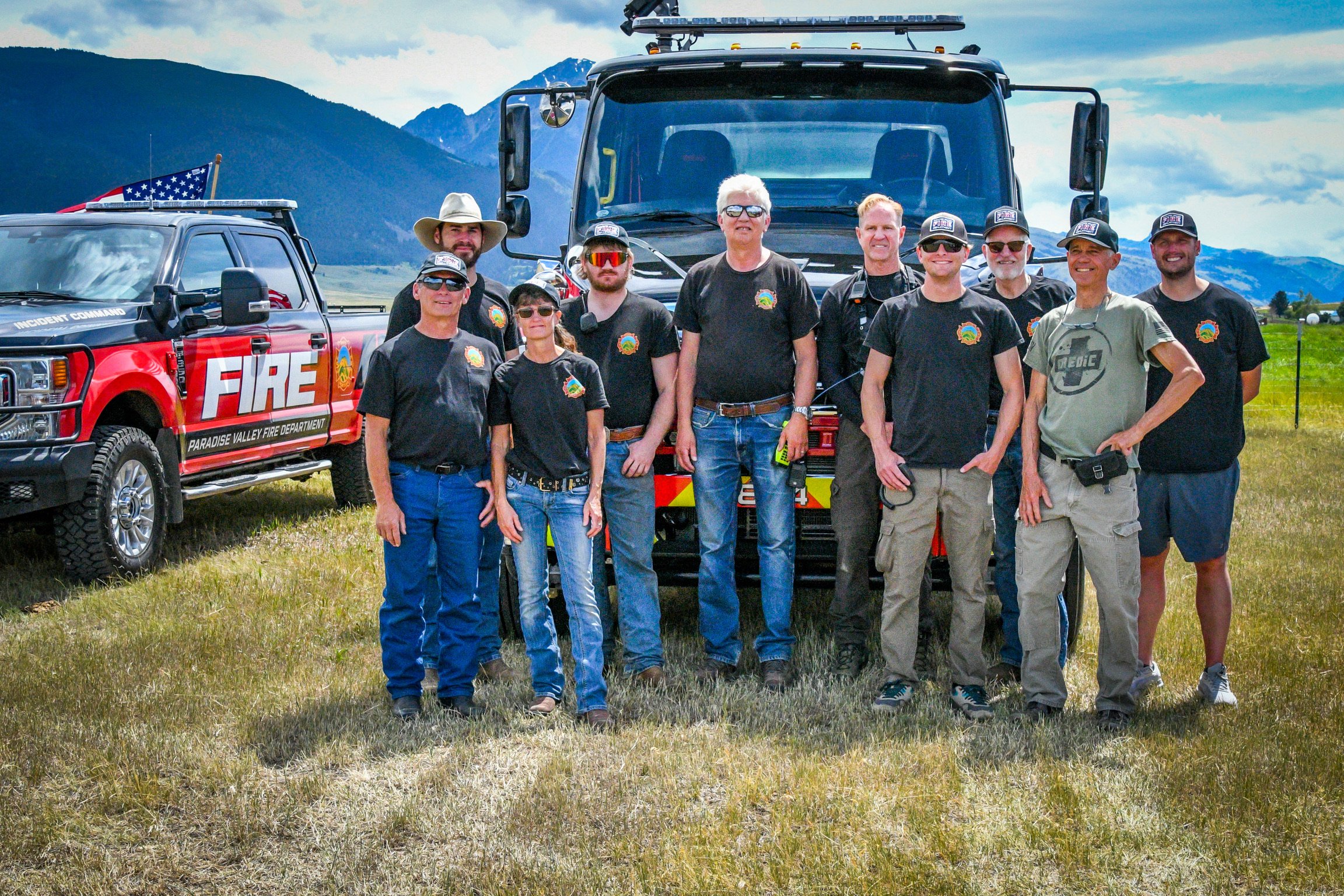 Group of firefighters standing on grassy field in front of fire trucks with mountain backdrop.