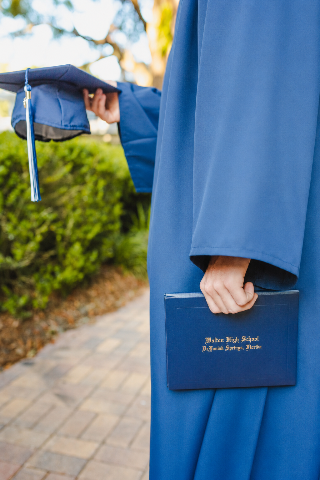 Close-up of a graduate in a blue gown holding a diploma with text 'Walton High School, H.J. Foster Springs, Florida' in a graduation ceremony outdoors.