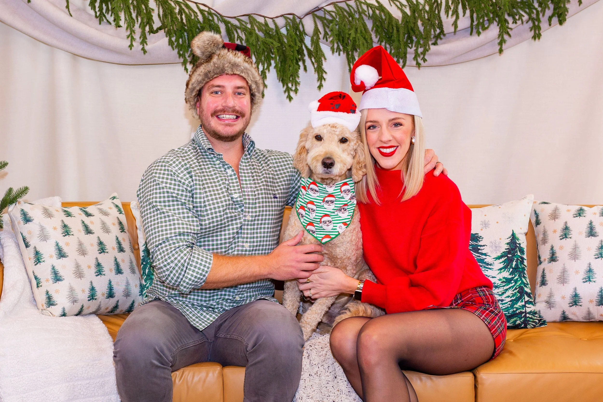 A cheerful couple and their dog sitting on a sofa decorated for Christmas with greenery overhead and pillows with forest designs, all wearing Christmas hats.