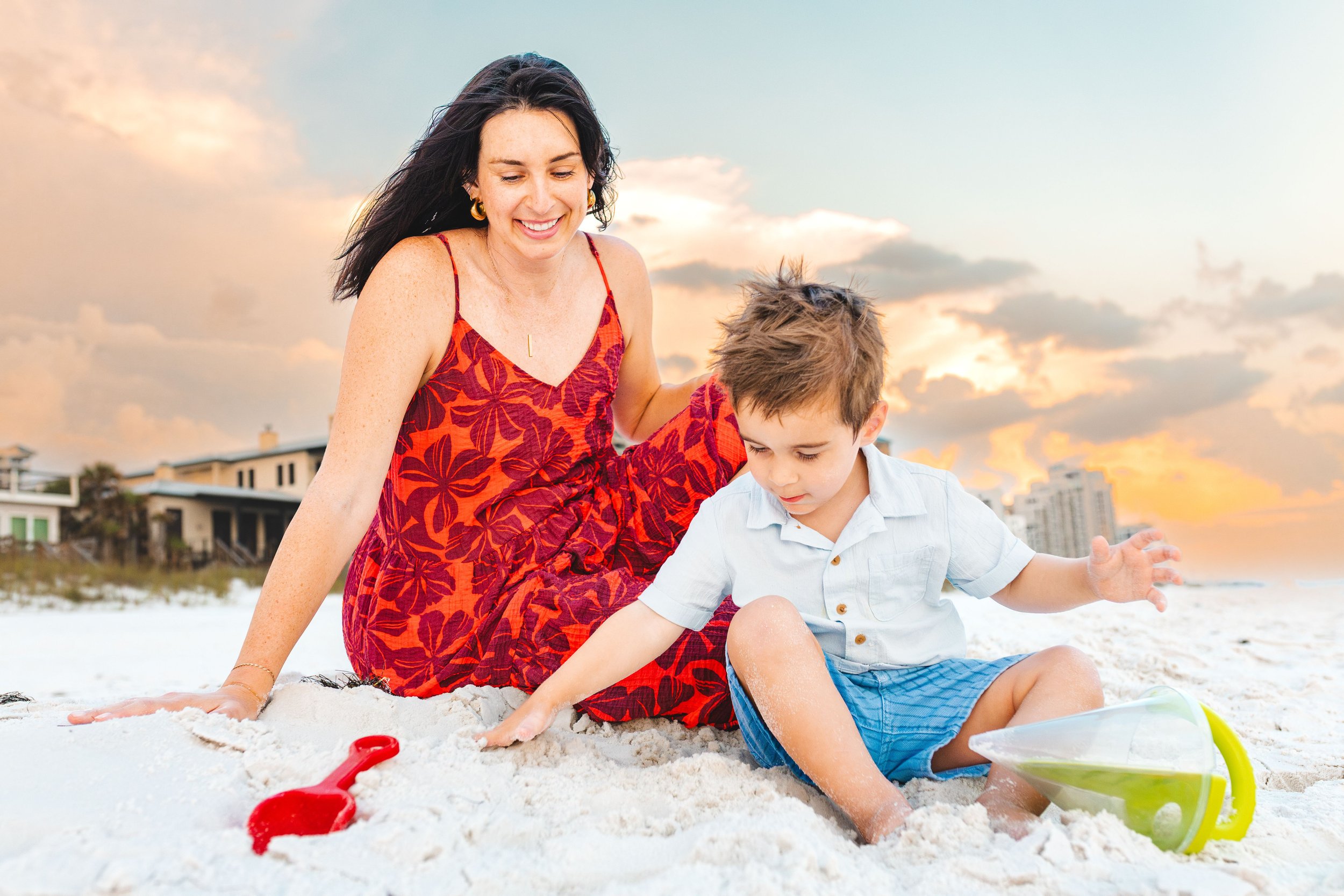 A woman and a young boy playing in the sand on a beach at sunset, with residential buildings and a colorful sky in the background.