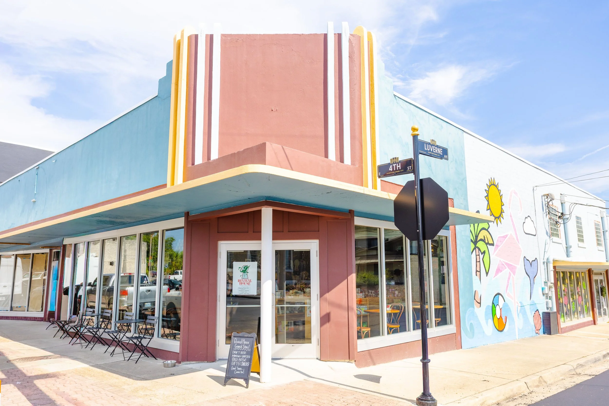 Colorful building with pastel pink, blue, and yellow exterior, with a mural of flowers and a sun on the wall. Outdoor seating with black chairs and a chalkboard sign in front, and street signs for 4th Street and Luverne Avenue.