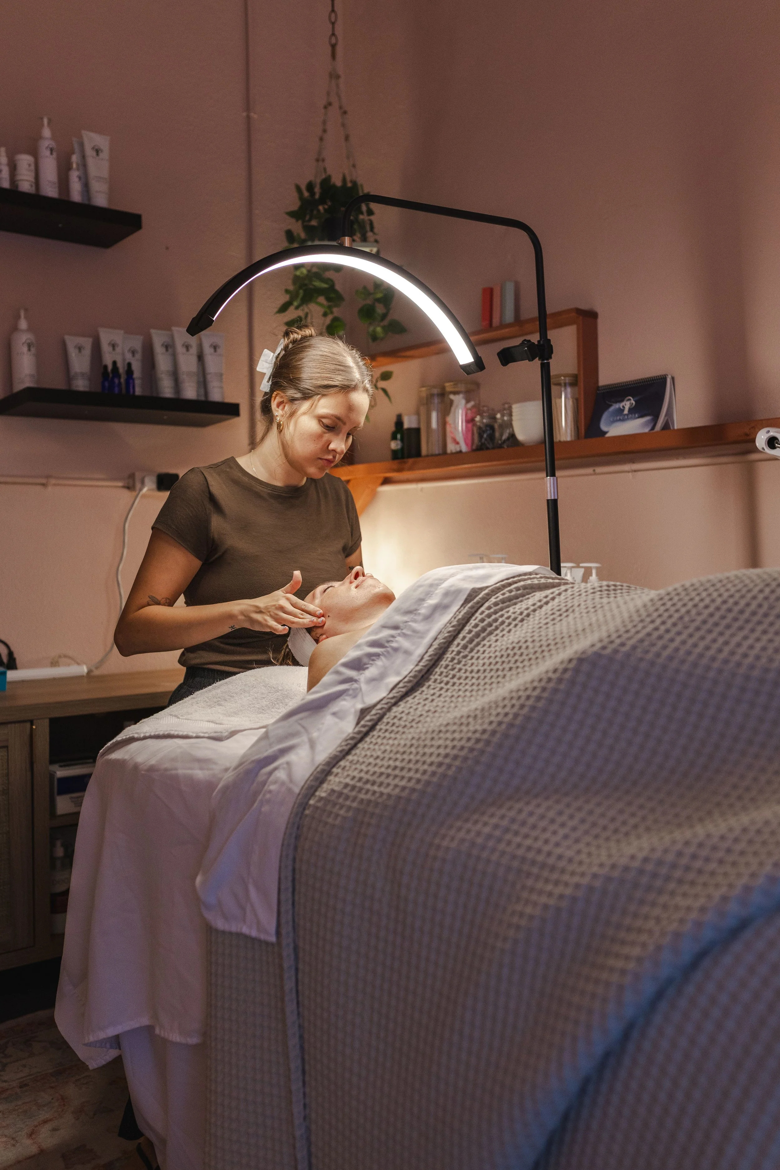 A woman receiving a facial treatment at a spa or wellness center, lying on a bed with a blanket, while a skincare professional applies a treatment under a bright, curved LED light.