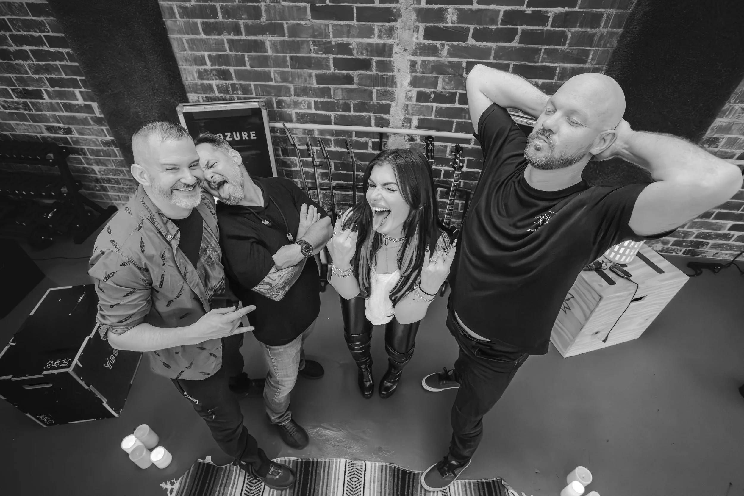 Four people posing for a photo in front of a brick wall, with two men and one woman making playful expressions, and one man smiling with his eyes closed, all standing close together. There are musical instruments and a sign that says 'AZURE' in the b