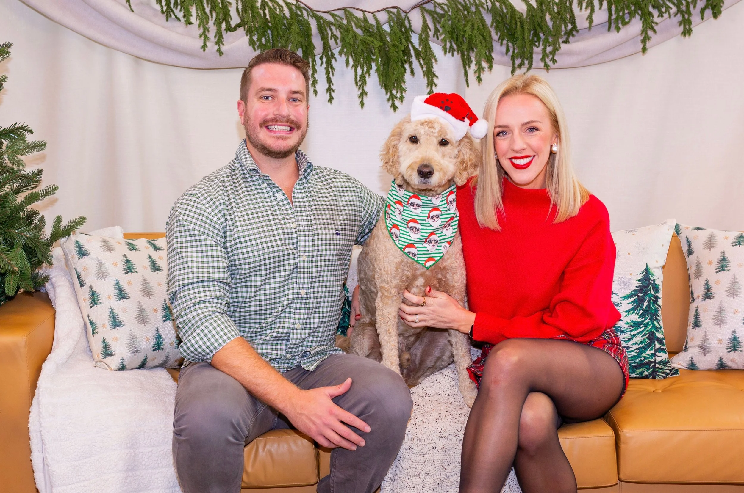 A man and woman sitting on a Christmas decorated couch with a dog wearing a Santa hat and holiday bandana between them. The woman is wearing a red sweater and plaid skirt, and the man is wearing a checkered shirt. There are Christmas trees and greene