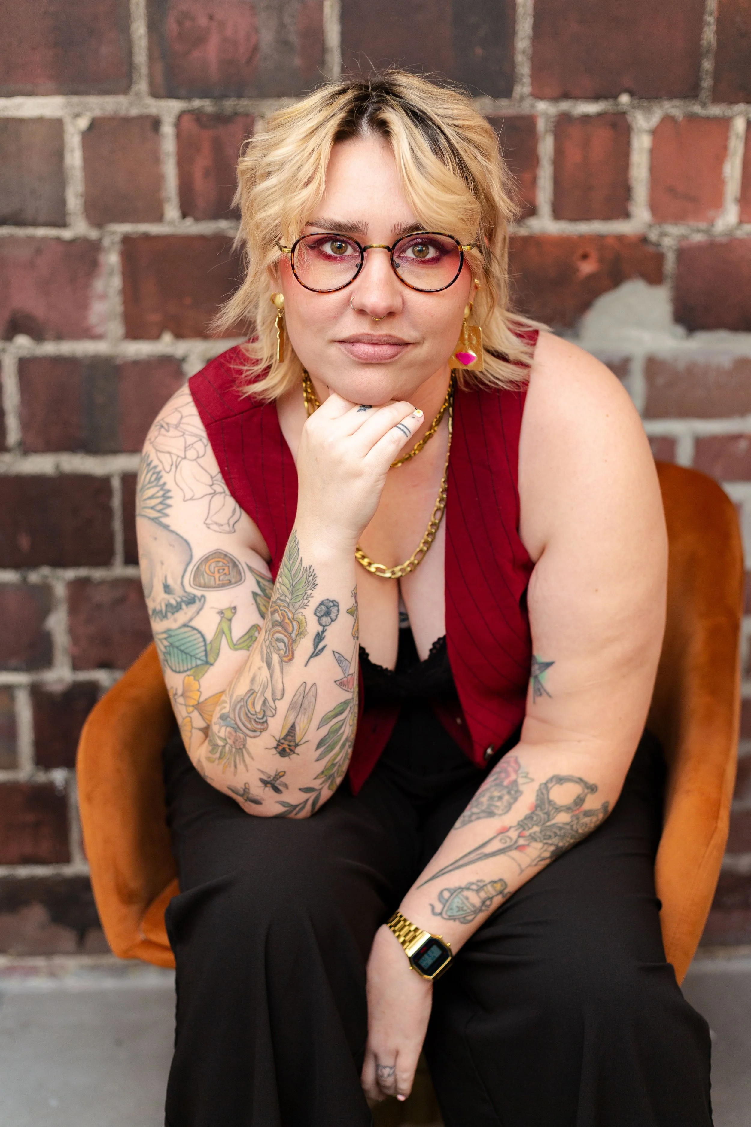 A young woman with short blonde hair wearing glasses, gold jewelry, and a sleeveless red top, sitting on a vintage brown armchair against a brick wall. She has tattoos on her left arm and right forearm, and its adorned with a gold watch on her left w