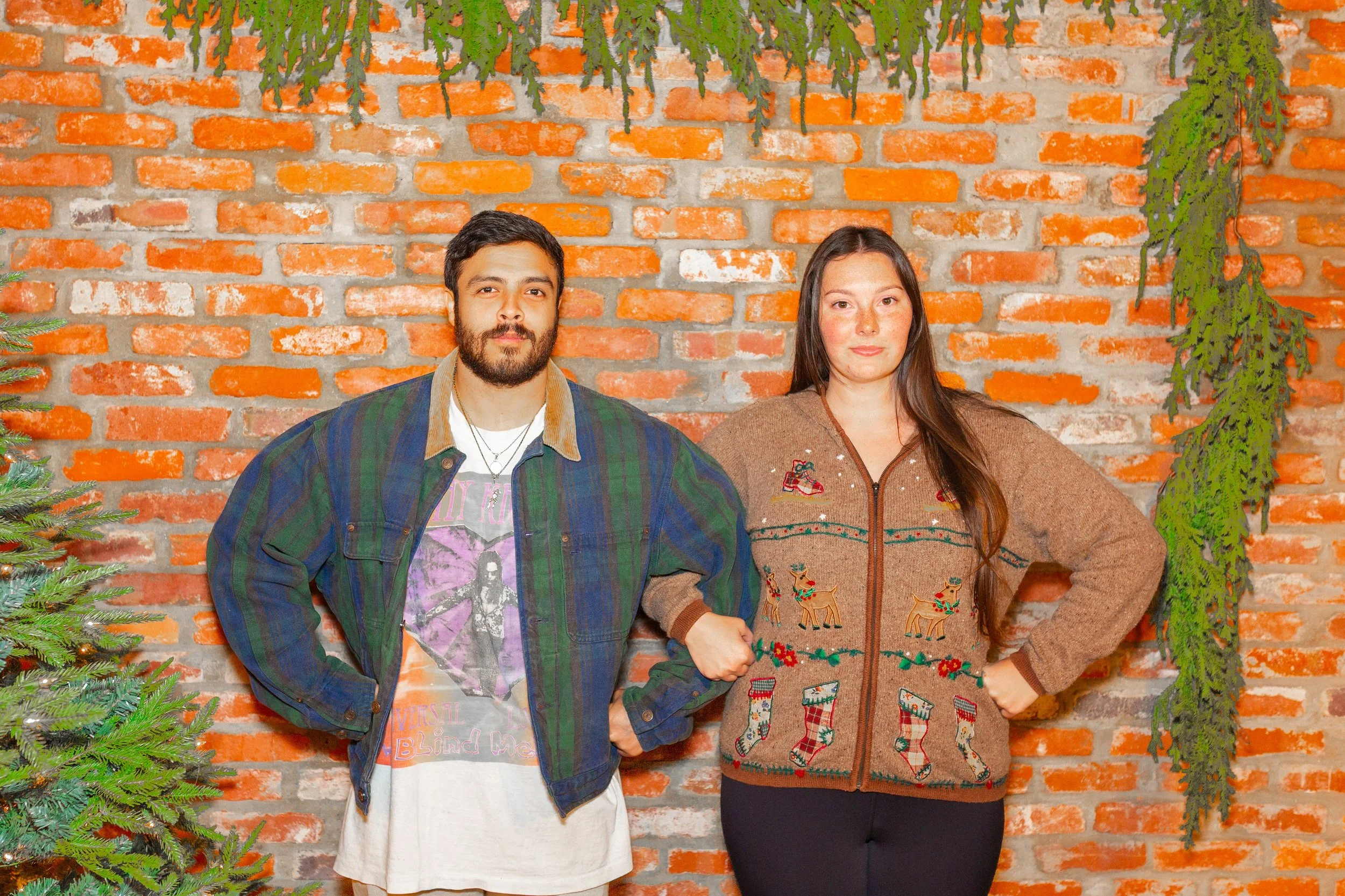 A man with a beard and a woman with long brown hair standing against a brick wall decorated with hanging greenery. The man is wearing a green plaid jacket over a graphic t-shirt, and the woman is wearing a brown Christmas sweater with holiday motifs 