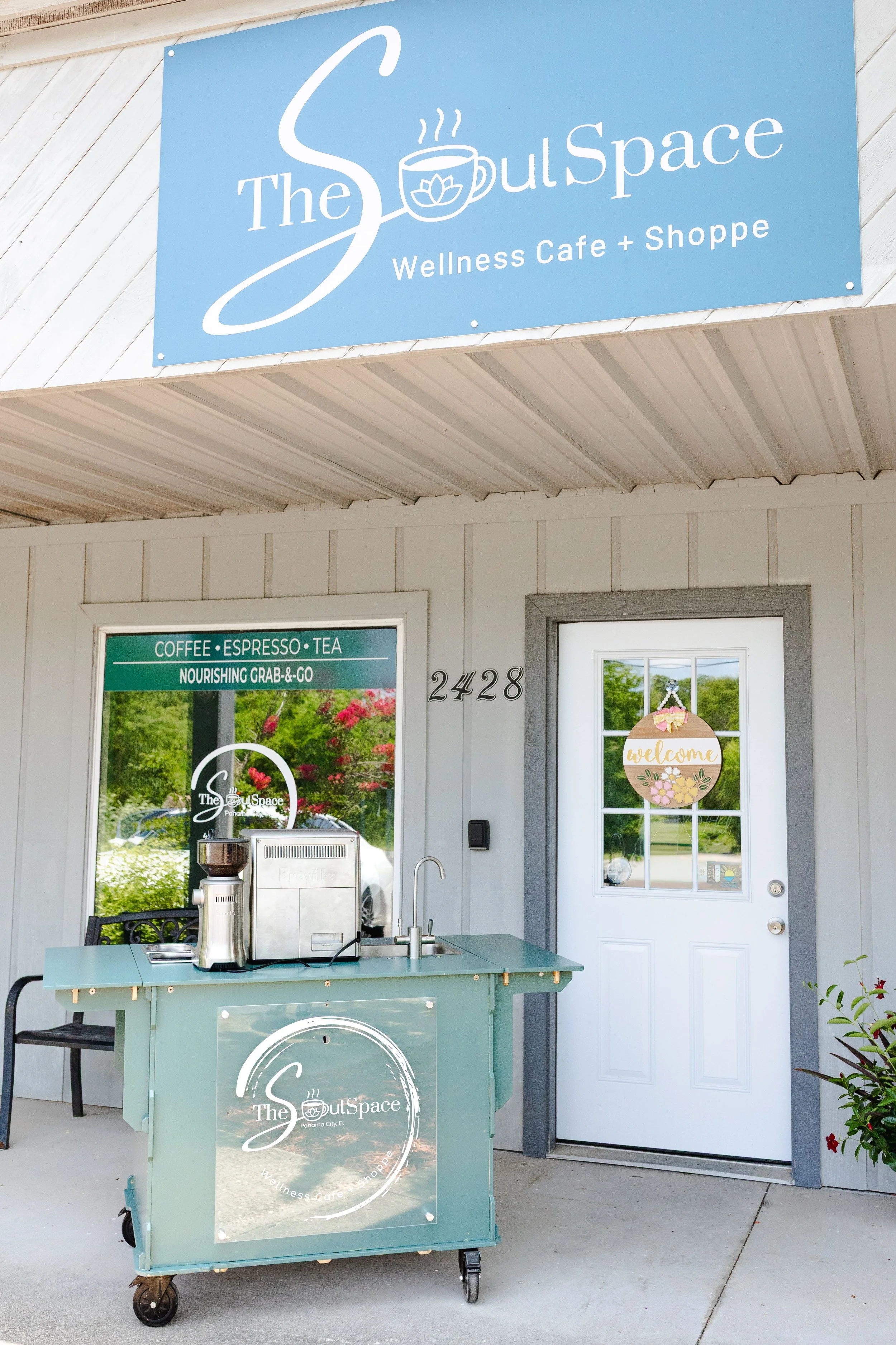 The exterior of The Soul Space Wellness Cafe and Shoppe, with a large blue sign displaying the name and logo, a decorated door with a welcome sign, and a light blue mobile cart with the cafe's logo parked outside.