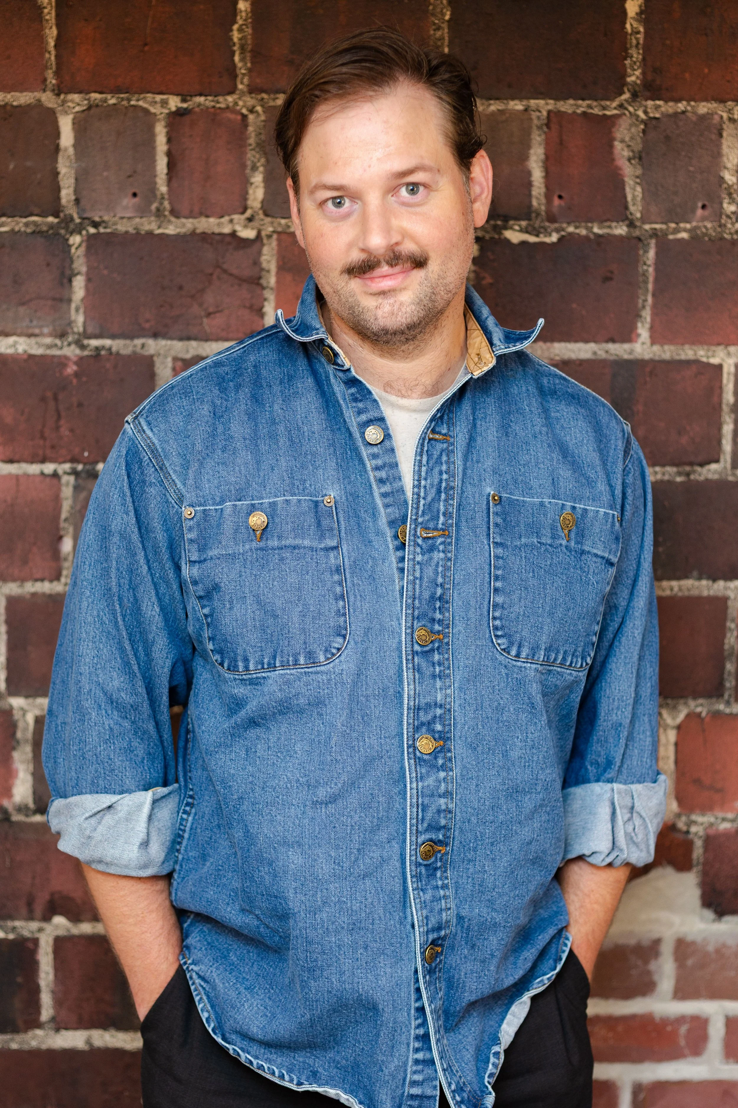 A man with light skin, light blue eyes, and brown hair with a mustache and beard, standing in front of a brick wall. He is wearing a blue denim shirt with rolled-up sleeves and has his hands in his pockets.