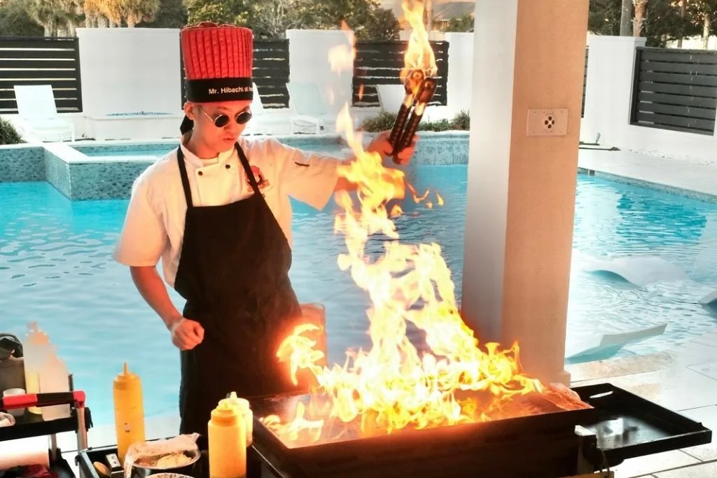 A chef wearing a tall red hat and sunglasses cooks at an outdoor grill with flames, next to a swimming pool.