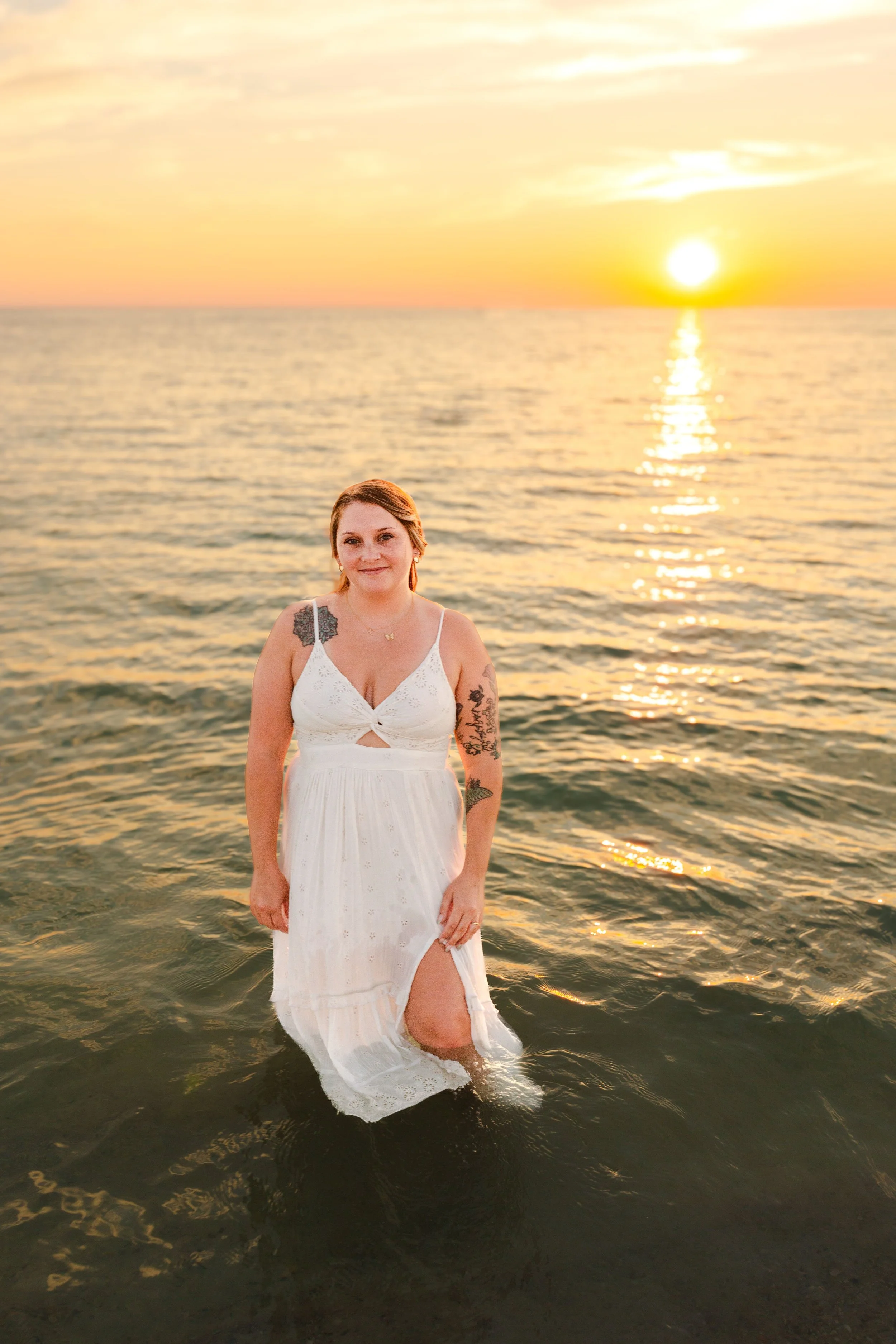 A woman standing in the ocean water at sunset, wearing a white dress with tattoos on her arms, with the sun setting over the horizon in the background.