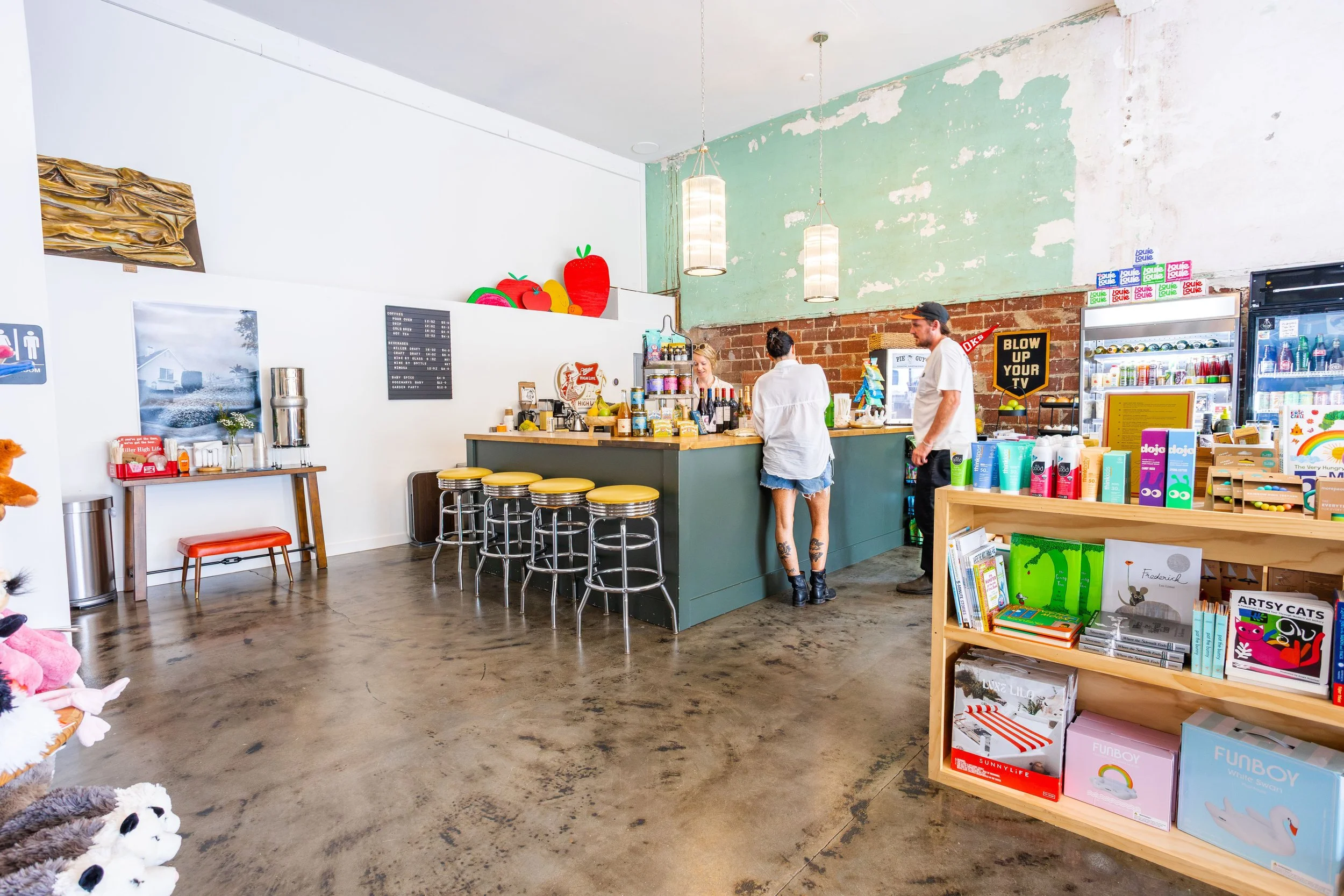 Inside a cafe or retail shop with a counter, bar stools, and shelves filled with books and toys. Two people are at the counter, with one behind it preparing drinks and the other waiting. Decor includes colorful fruit-shaped wall art, hanging lights, 