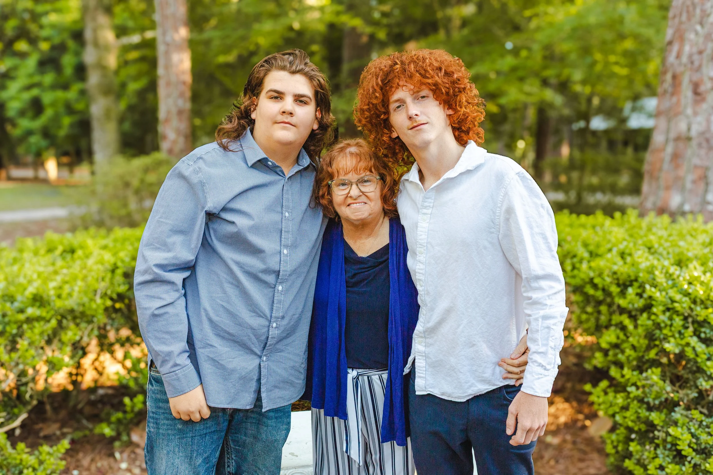 A woman with red curly hair and glasses standing between two young men with curly and straight hair, all outdoors in a wooded area.
