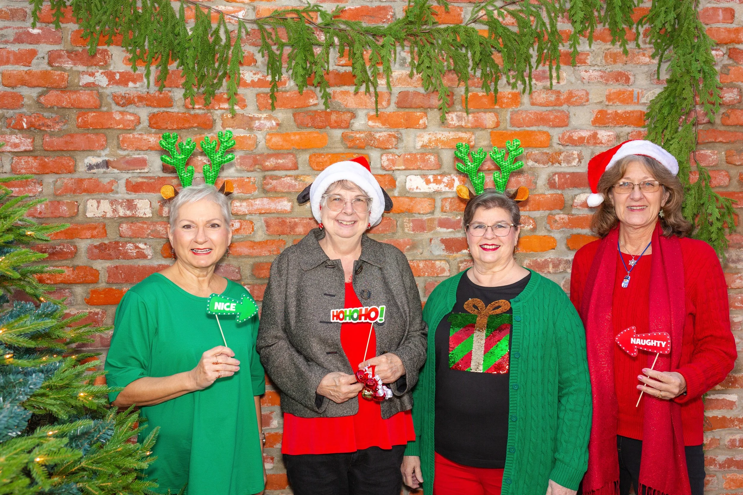 Four women dressed in Christmas sweaters and hats posing in front of a brick wall decorated with green foliage, with some holding holiday-themed props and signs.