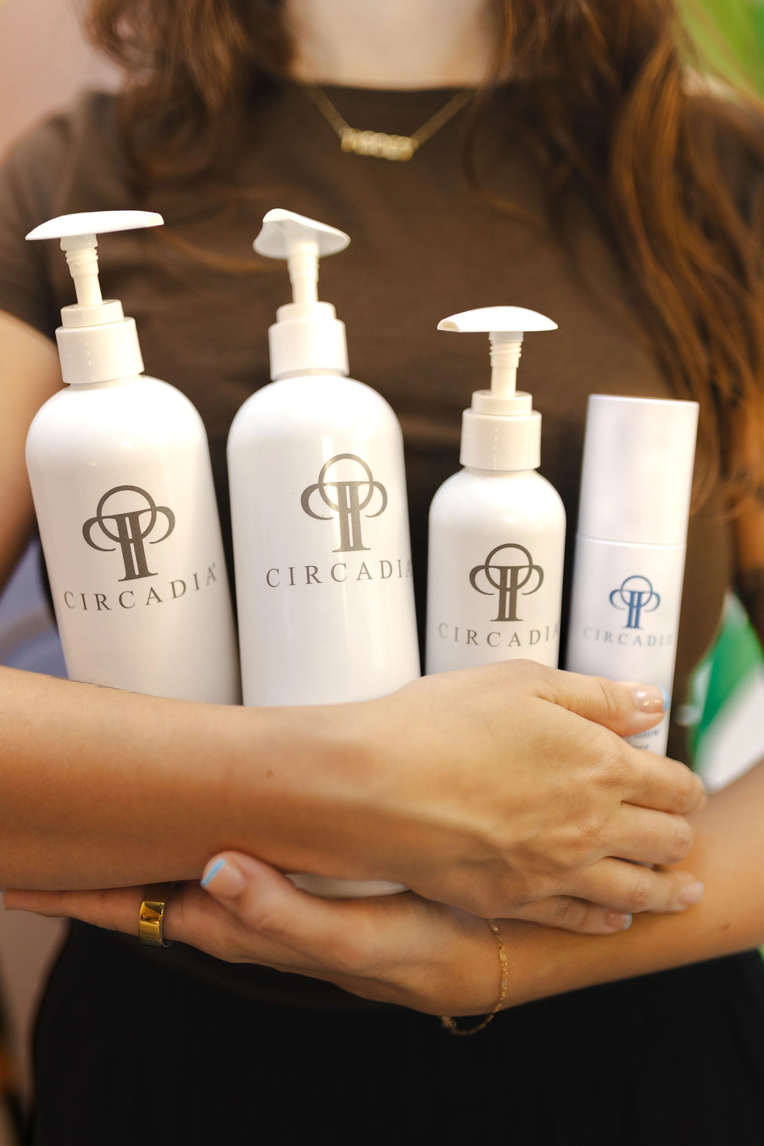 Person holding four white skincare bottles with pump dispensers labeled 'CIRCADIA' and a small section of their torso showing. The person is wearing a black top, gold jewelry, and has long, wavy buckskin-colored hair.