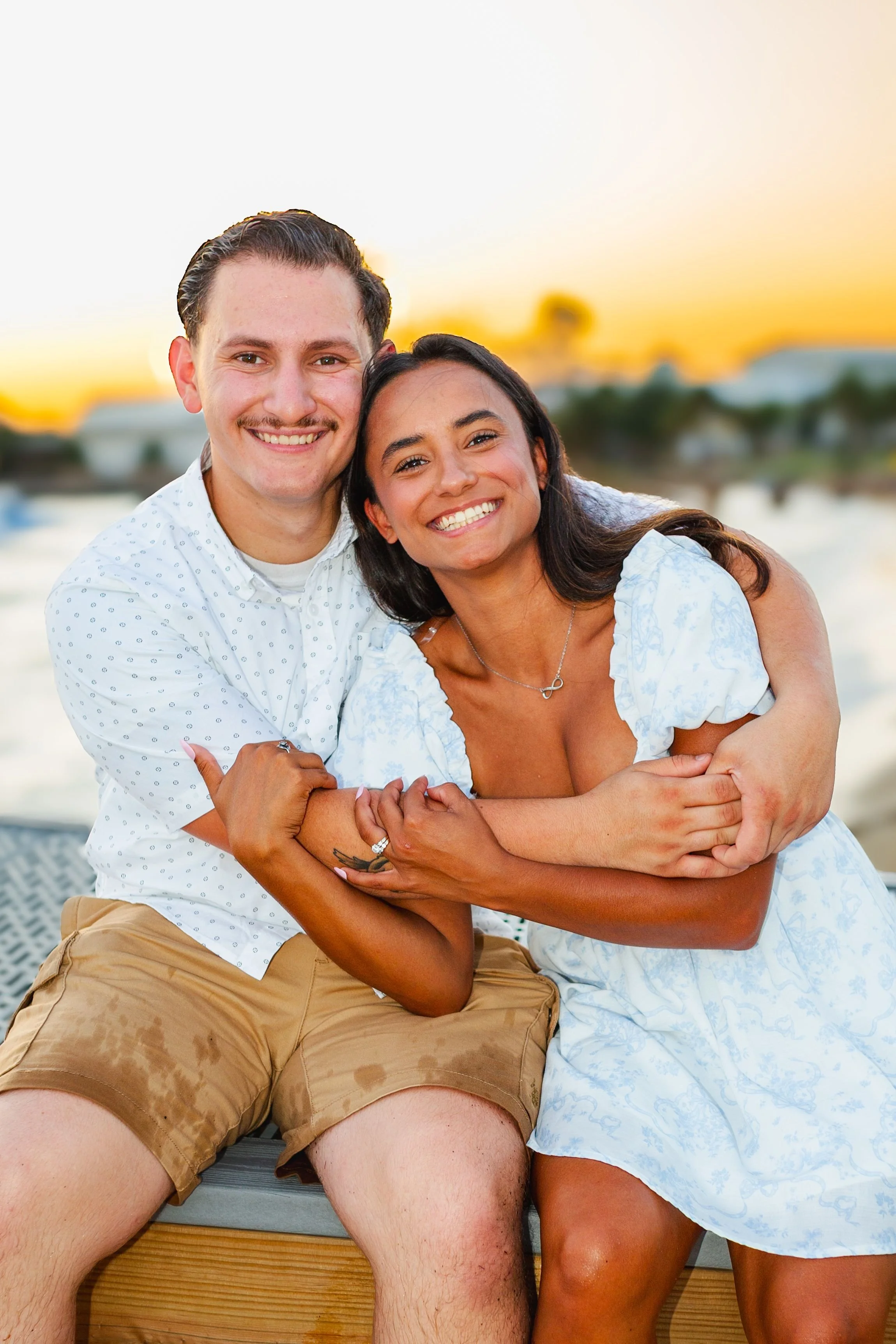A smiling man and woman sitting close together outdoors during sunset, with the woman embracing the man. The background features a blurred landscape with water and trees.
