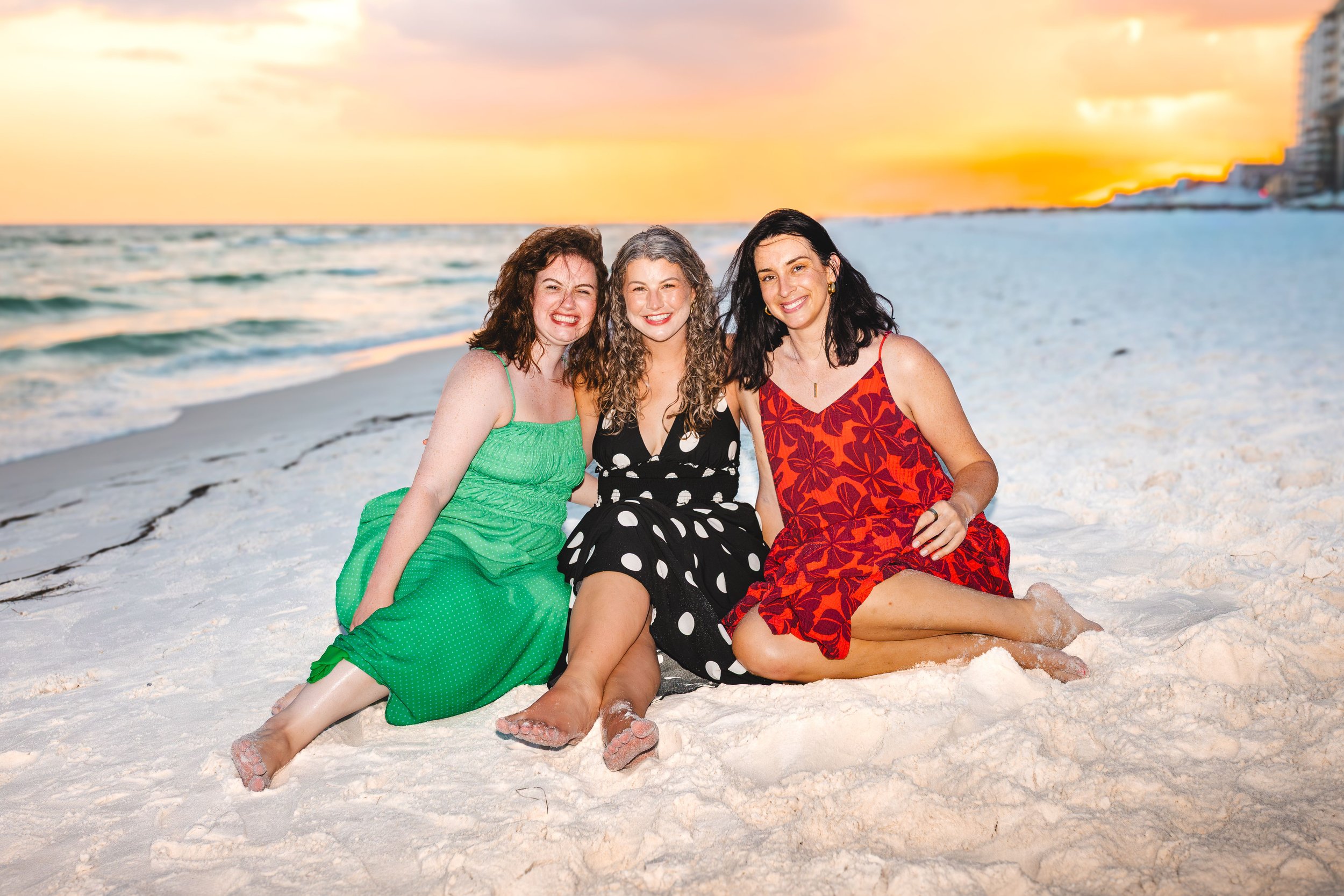 Three women sitting on the sandy beach at sunset, smiling and enjoying their time together.