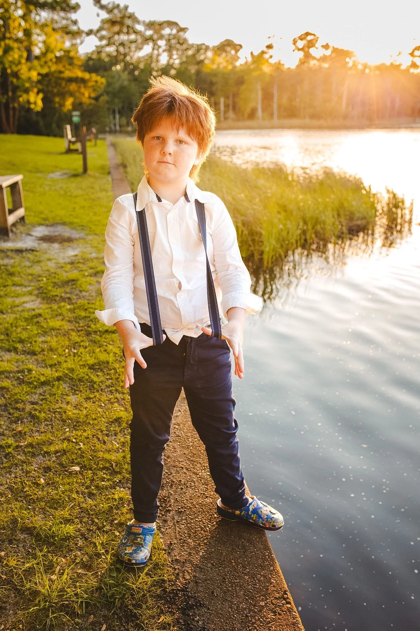 A boy with reddish hair wearing a white shirt, suspenders, dark pants, and colorful sneakers standing on a lakeside path during sunset.