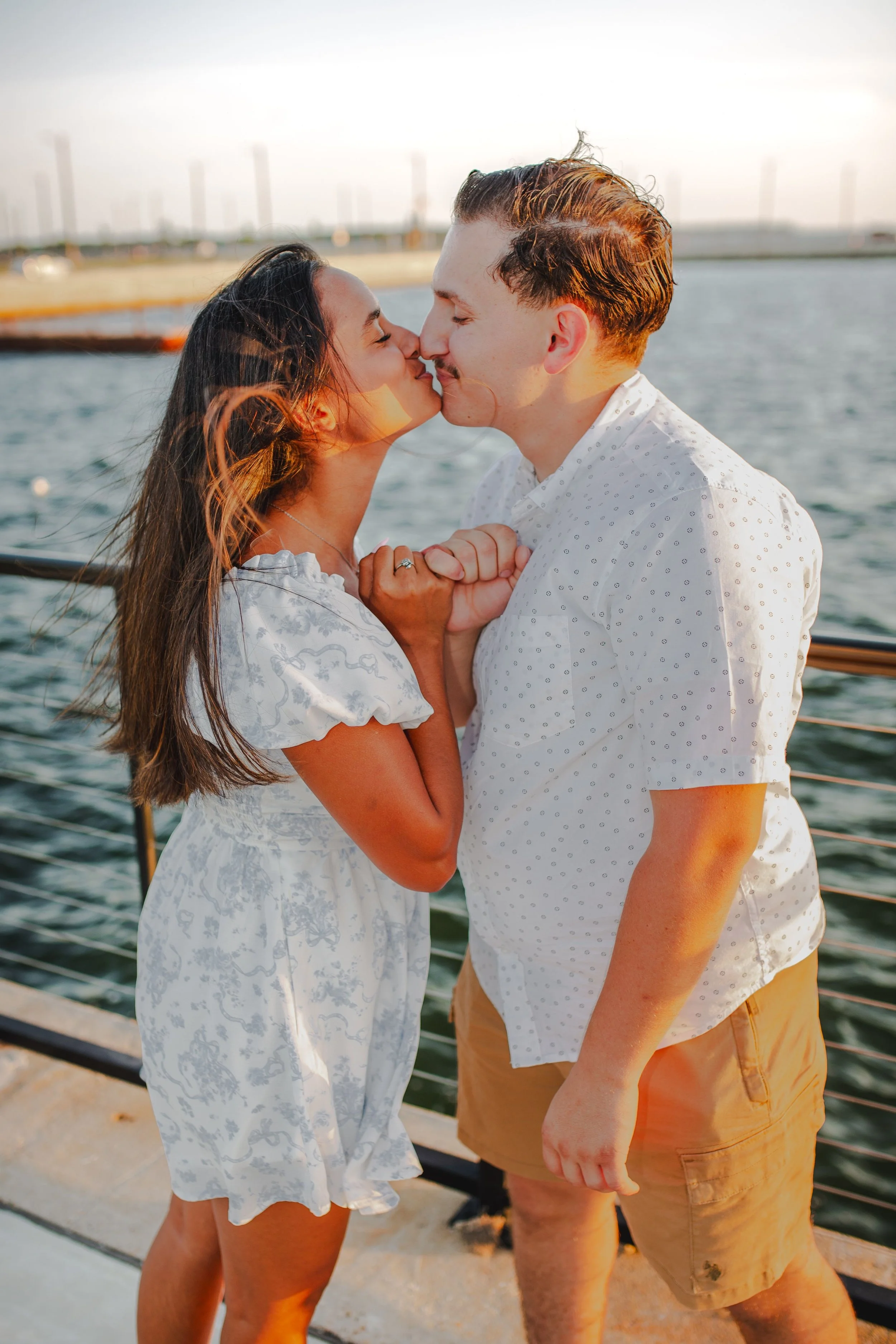 A couple is kissing by a waterfront during sunset, holding hands, with wind blowing through their hair, dressed in light summer clothing.