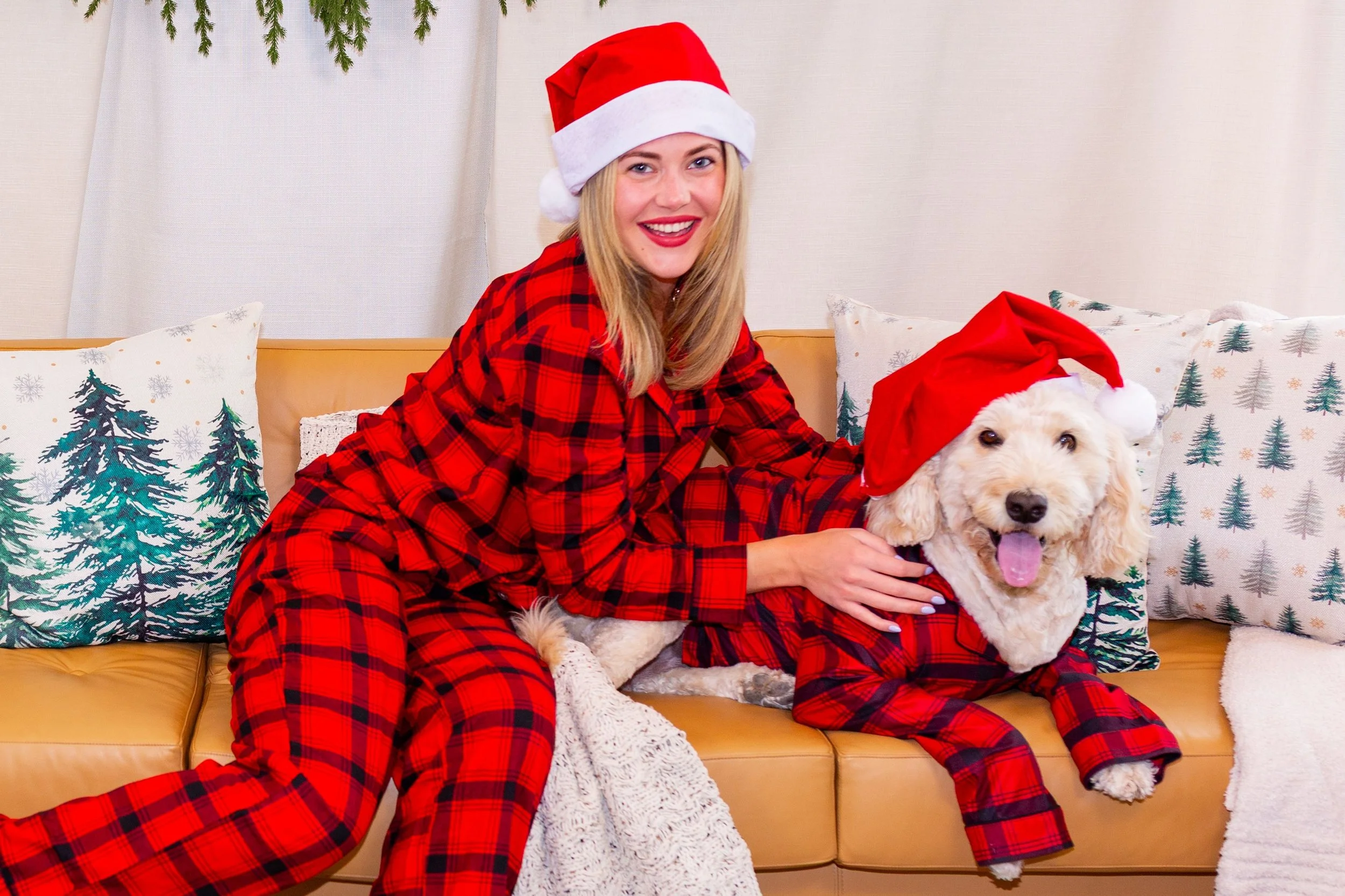 A woman dressed in red plaid pajamas and a Santa hat smiling while sitting on a yellow couch with a white dog wearing a Santa hat. The background has Christmas-themed pillows and a white curtain with greenery hanging above.