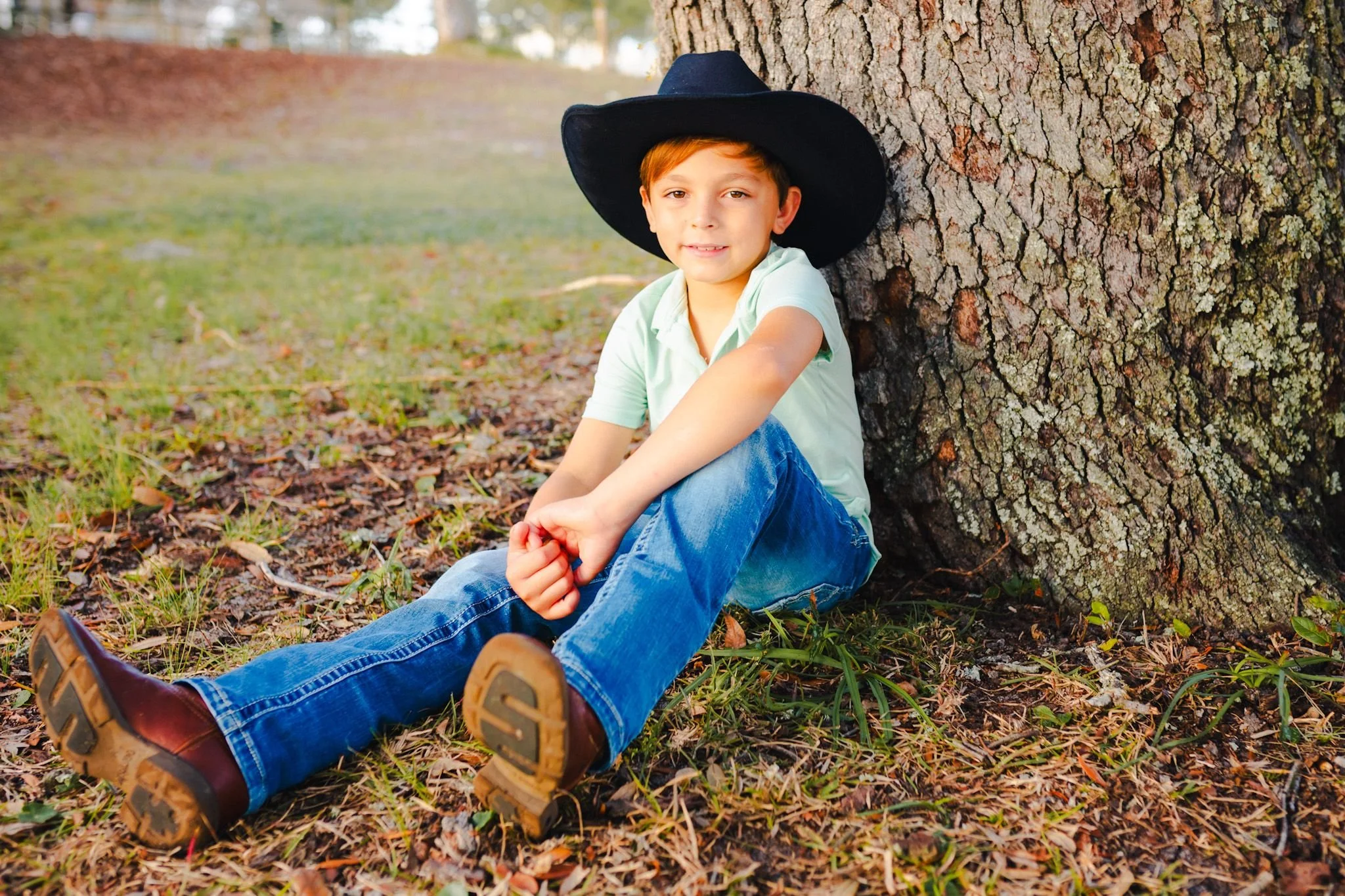 A young boy with red hair sitting on the ground next to a large tree trunk, wearing a black hat, light green shirt, blue jeans, and brown shoes, in a park with fallen leaves.