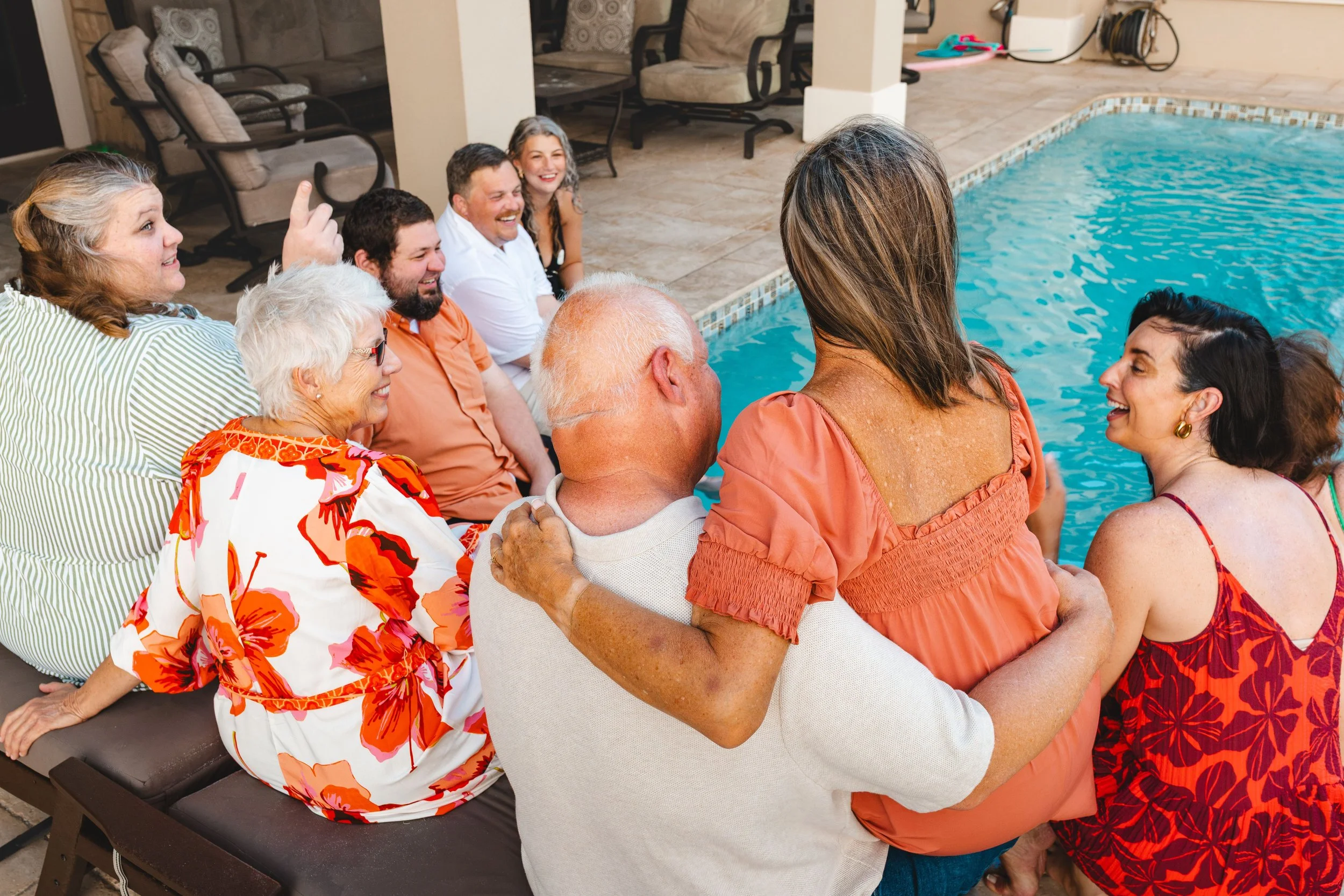 A group of people sitting and standing around a swimming pool, smiling and enjoying each other's company.