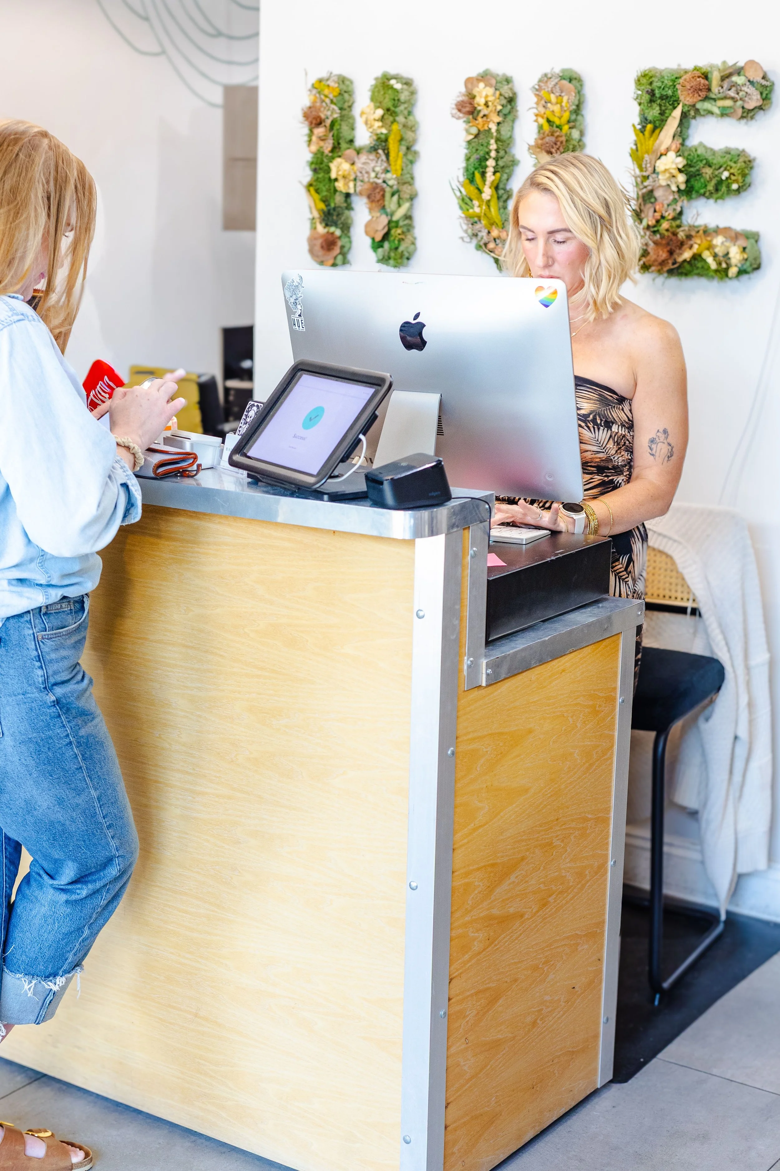 A woman with blonde hair working at a front desk with a computer, while another person is making a payment at the counter. The background has a wall with green and beige floral decor spelling out 'LOVE'.