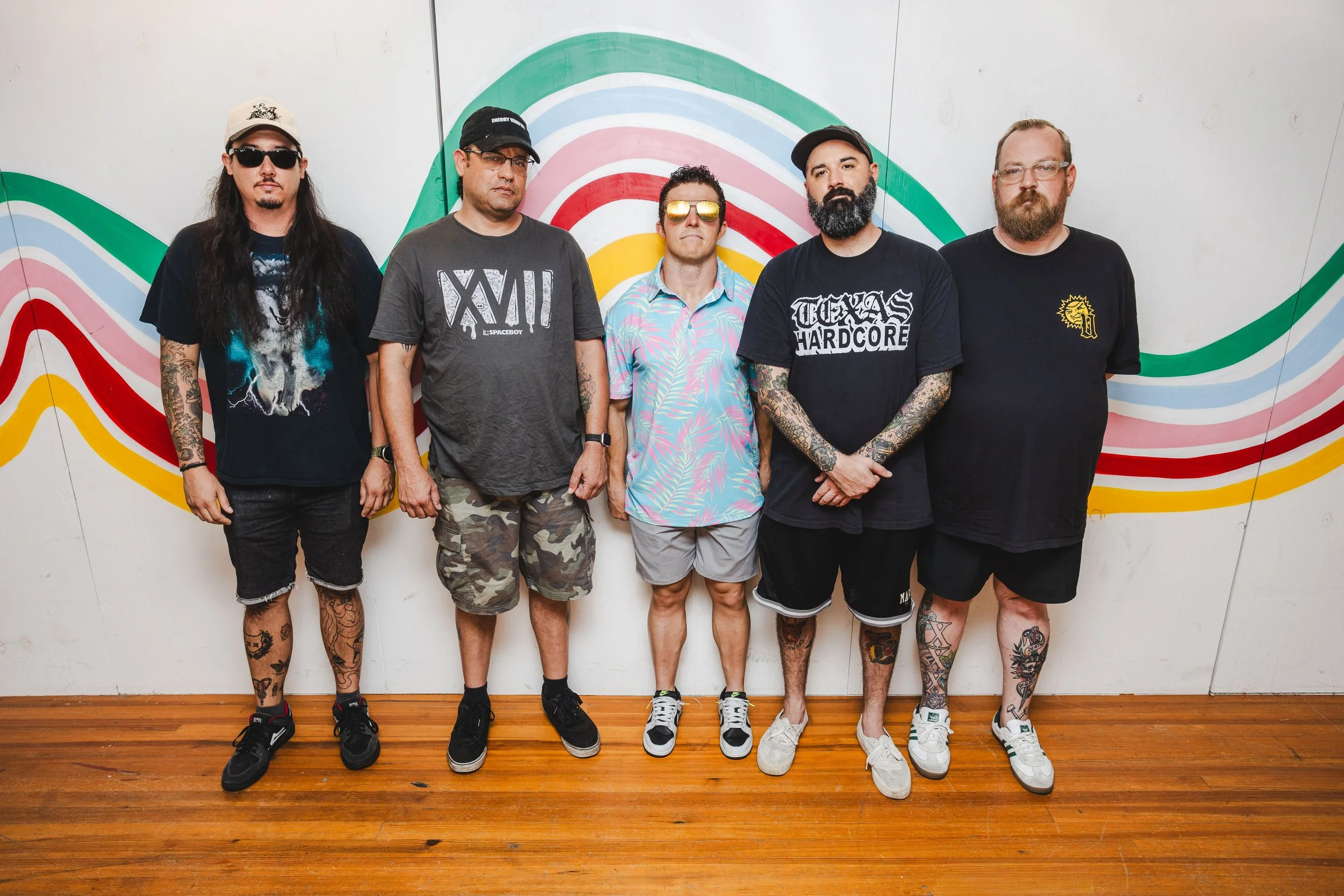 Group of five men standing indoors in front of a colorful, rainbow-patterned wall mural, wearing casual summer clothing, some with tattoos, sunglasses, and hats.