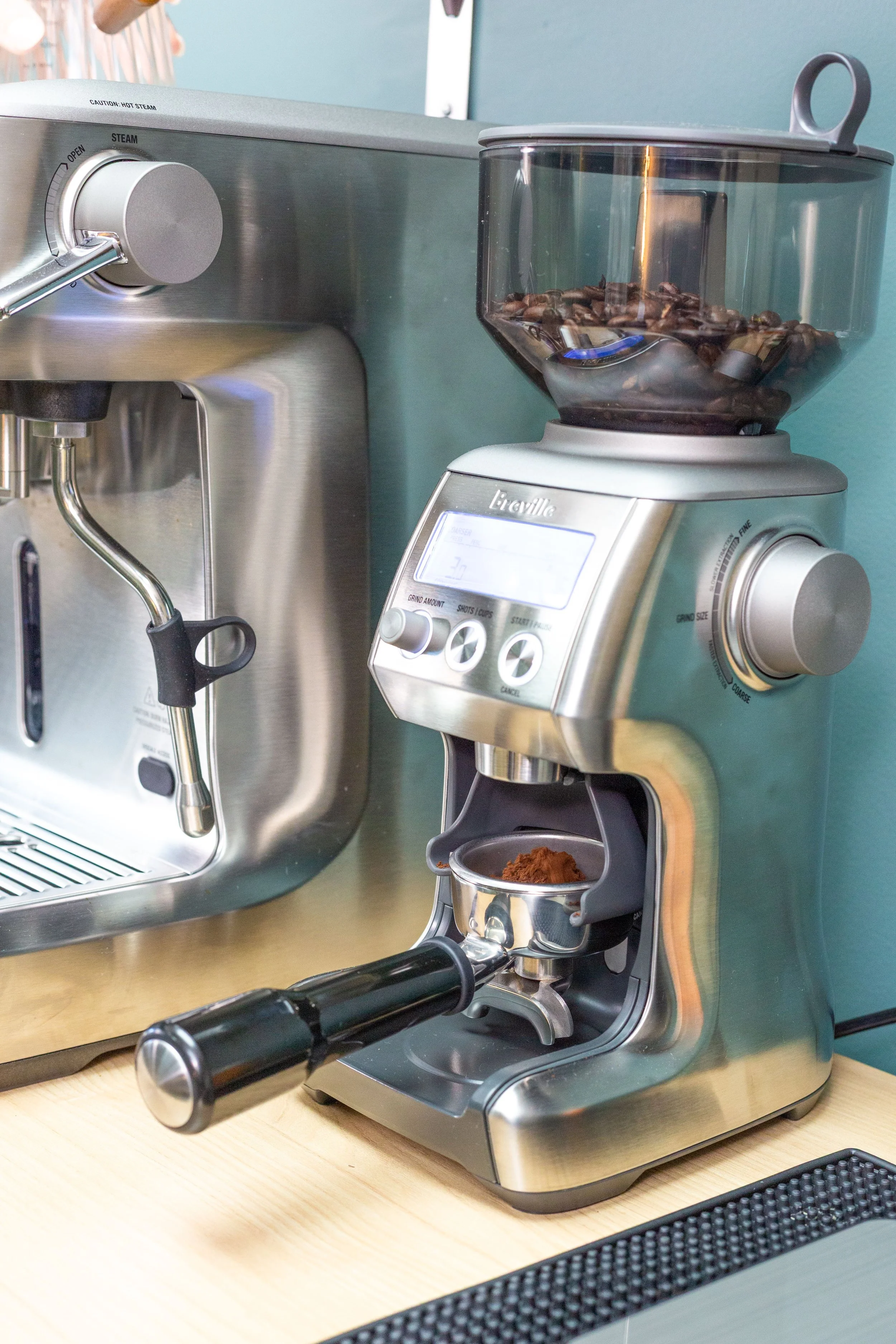 A coffee grinder with coffee beans inside, placed on a wooden surface. An espresso machine is visible in the background.
